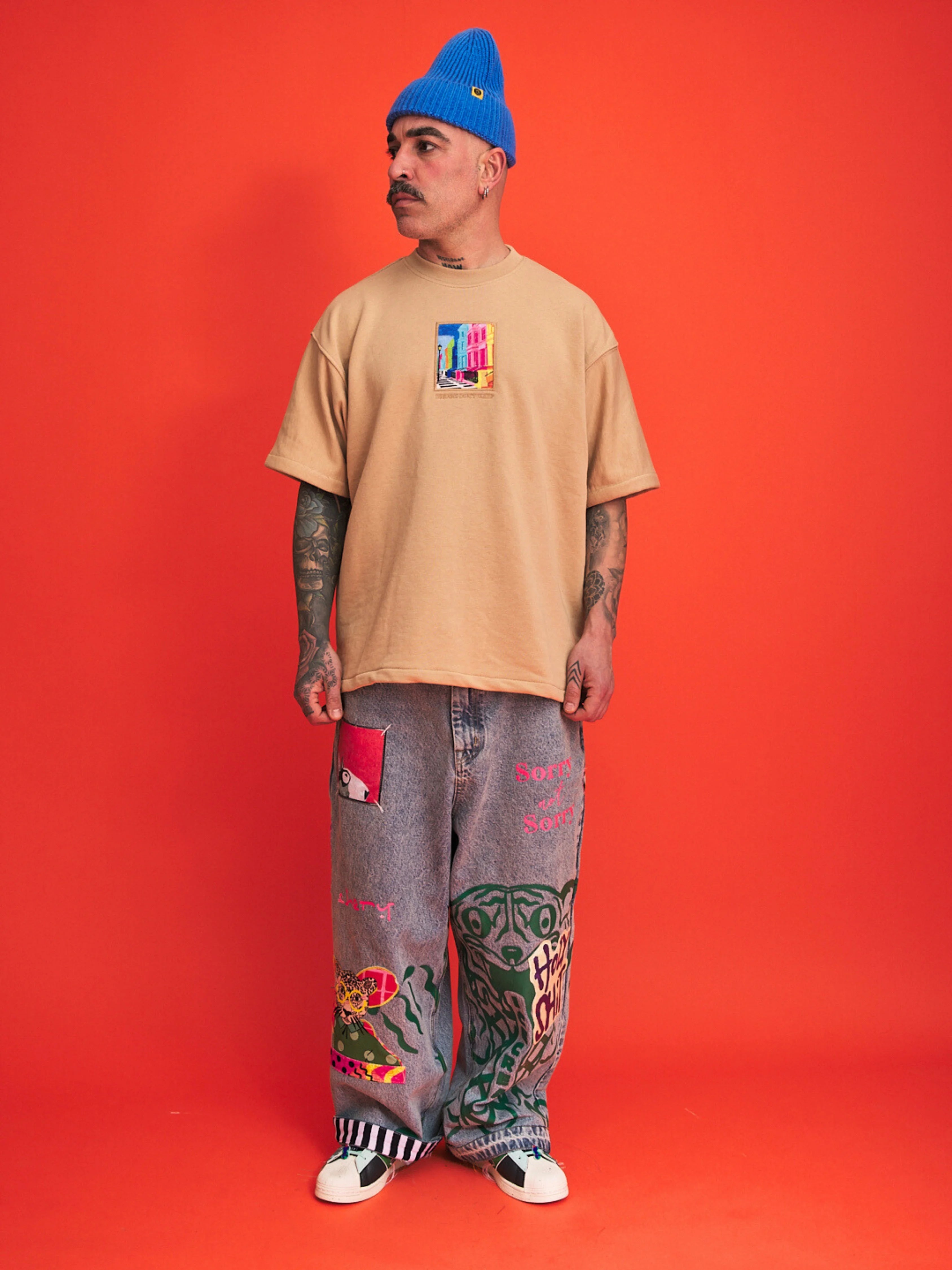 Person wearing a beige t-shirt, gray pants with designs, and a blue beanie against an orange background