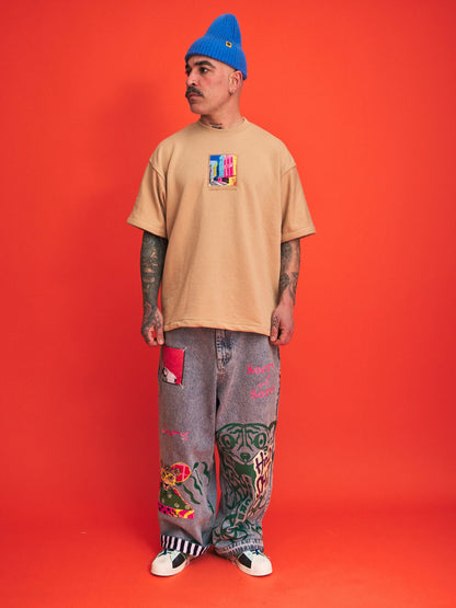 Person wearing a beige t-shirt, gray pants with designs, and a blue beanie against an orange background