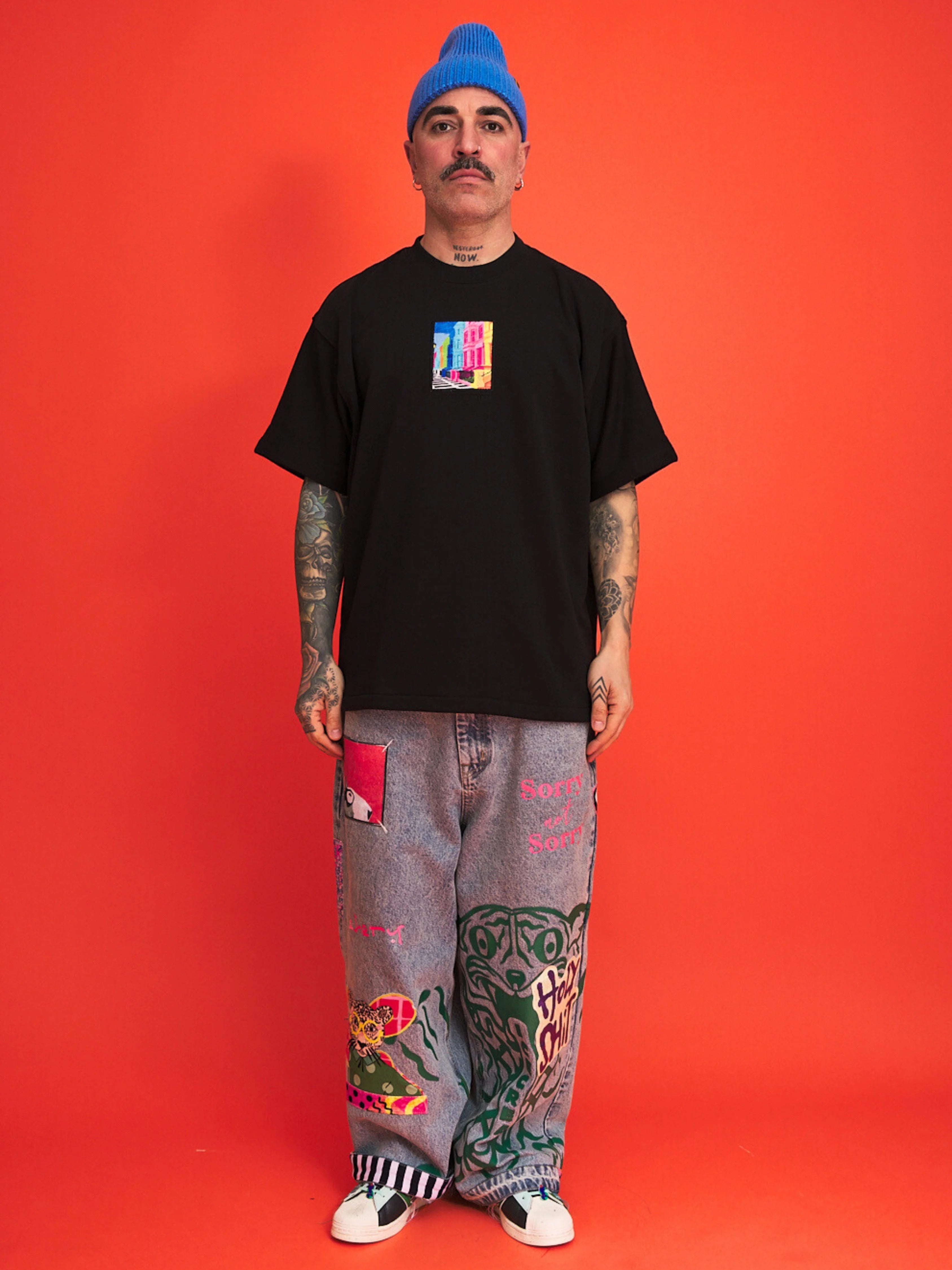 Person wearing a black t-shirt with a colorful graphic, gray pants with designs, and a blue beanie on an orange background.
