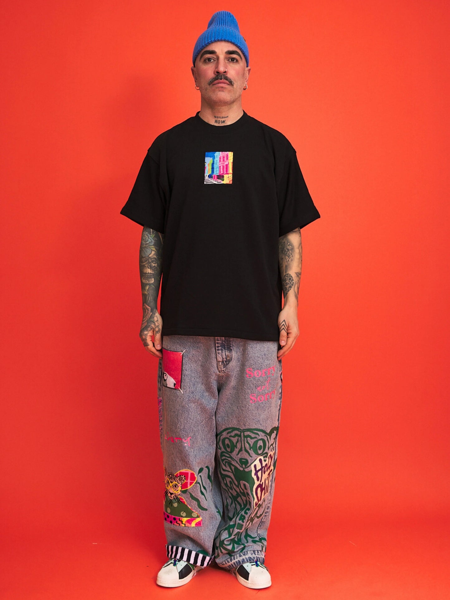 Person wearing a black t-shirt with a colorful graphic, gray pants with designs, and a blue beanie on an orange background.