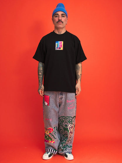 Person wearing a black t-shirt with a colorful graphic, gray pants with designs, and a blue beanie on an orange background.