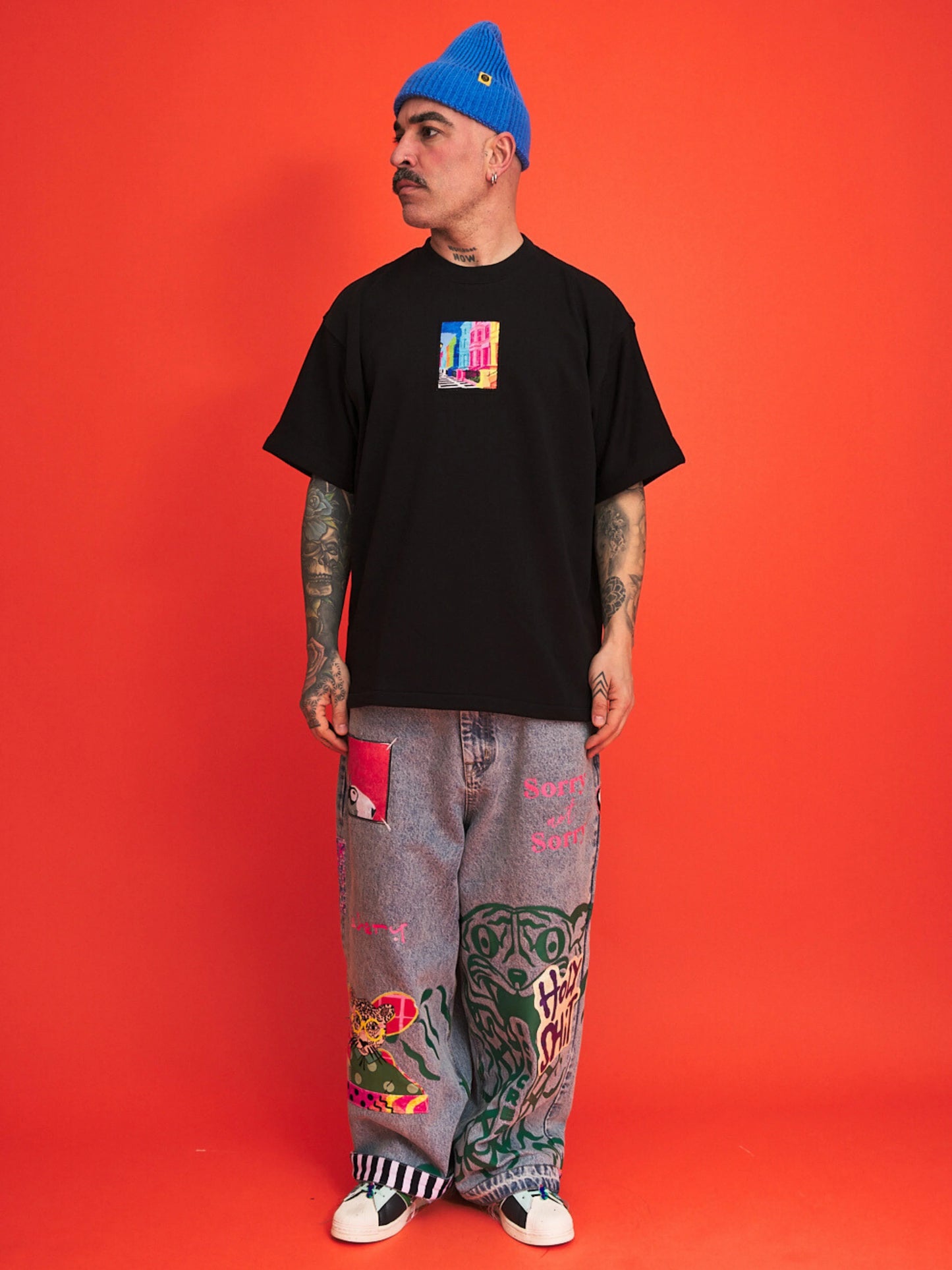 Person wearing a black t-shirt with colorful design, gray pants with patterns, and a blue beanie against an orange background