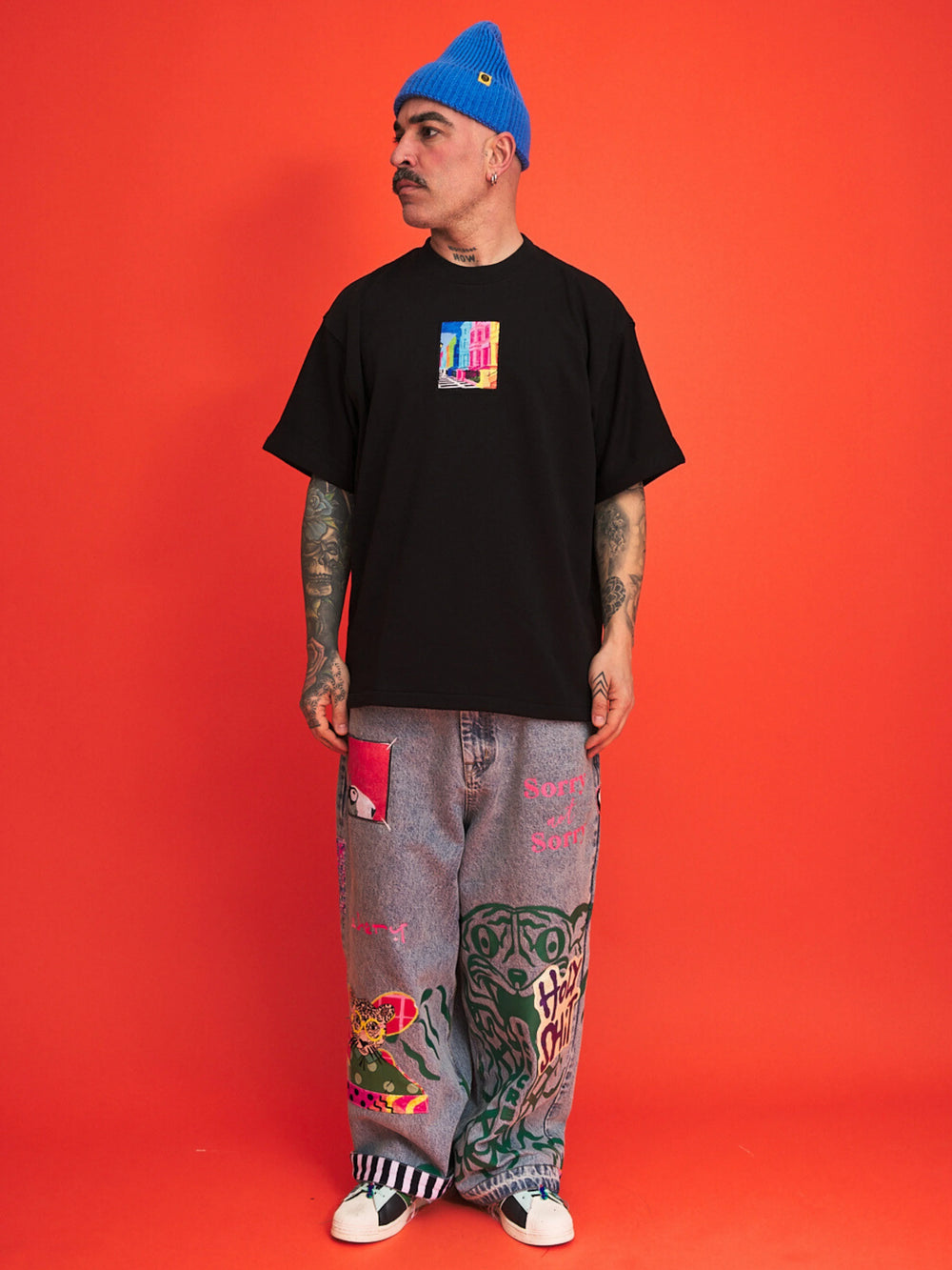 Person wearing a black t-shirt with colorful design, gray pants with patterns, and a blue beanie against an orange background