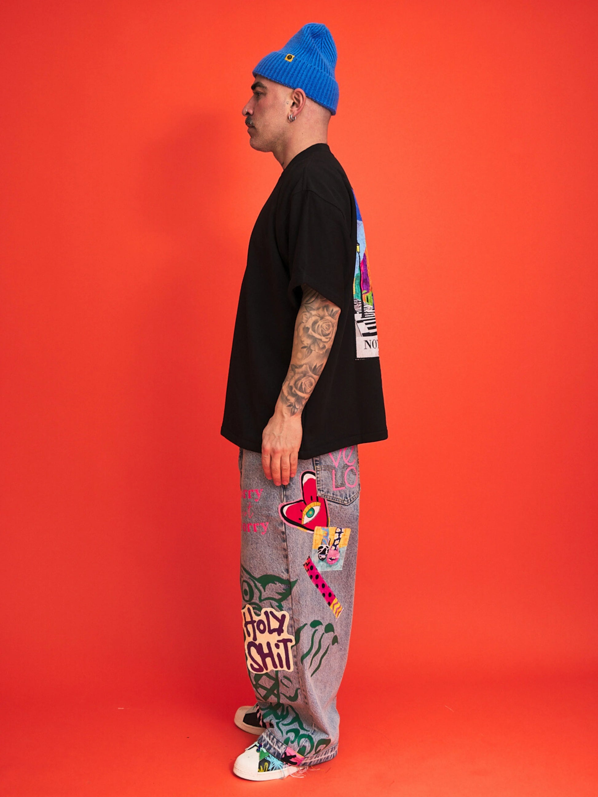 Person wearing a black t-shirt, colorful pants, and a blue beanie against an orange background