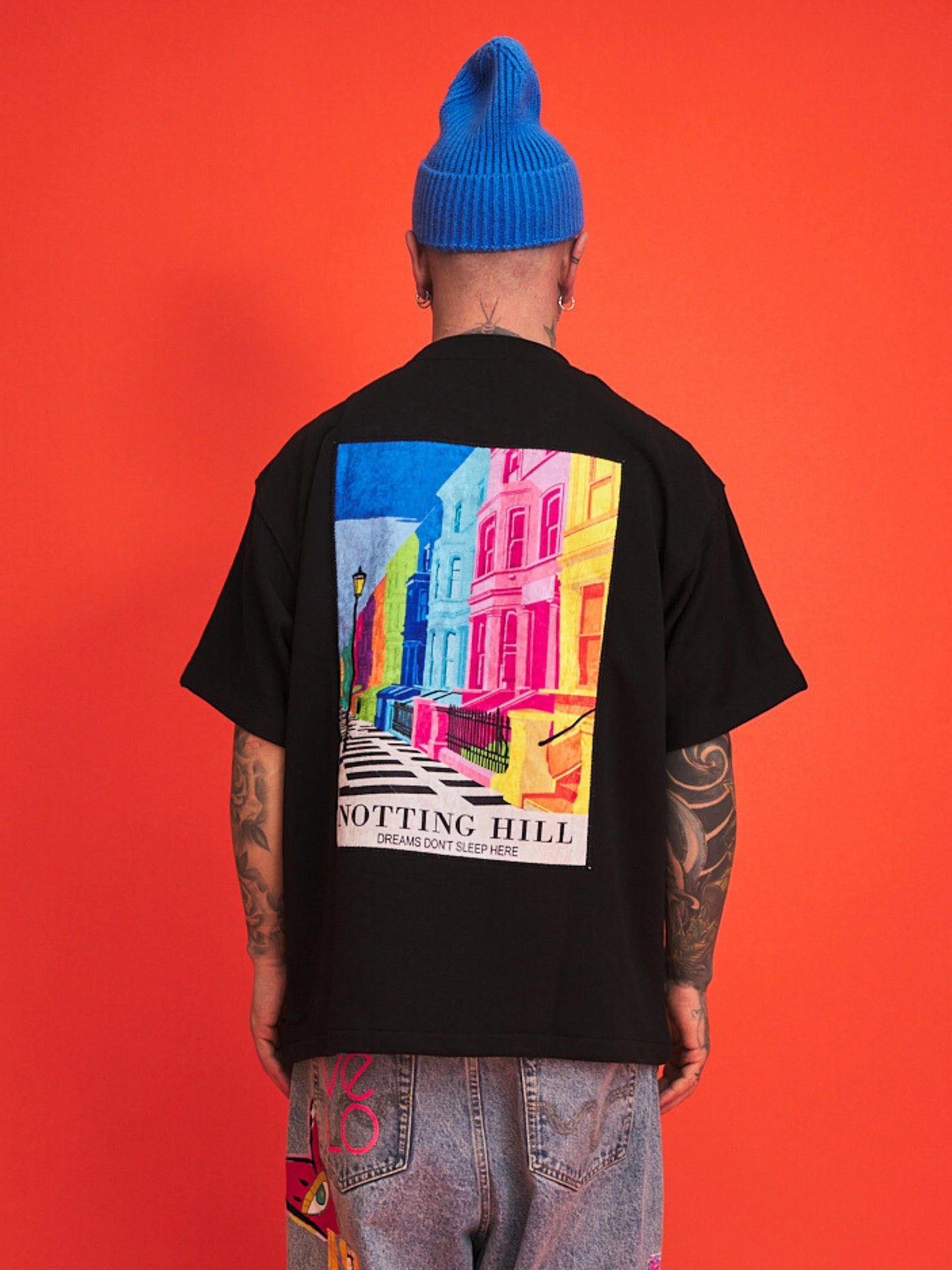 Person wearing a black t-shirt with a colorful 'Notting Hill' graphic on the back against an orange background.