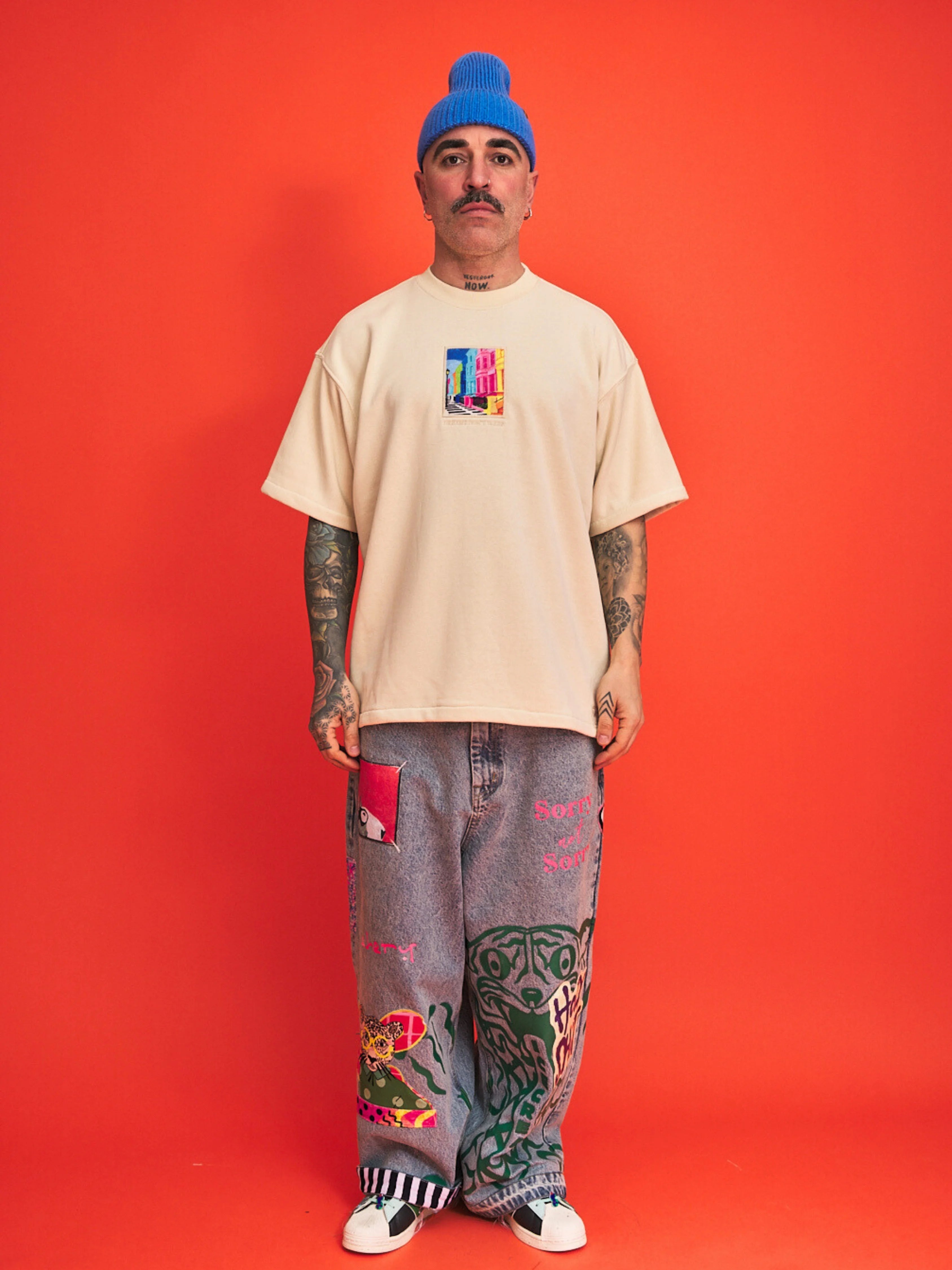 Person wearing a cream t-shirt with colorful graphic, gray pants with designs, and a blue beanie against an orange background