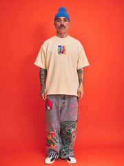 Person wearing a cream t-shirt with colorful graphic, gray pants with designs, and a blue beanie against an orange background