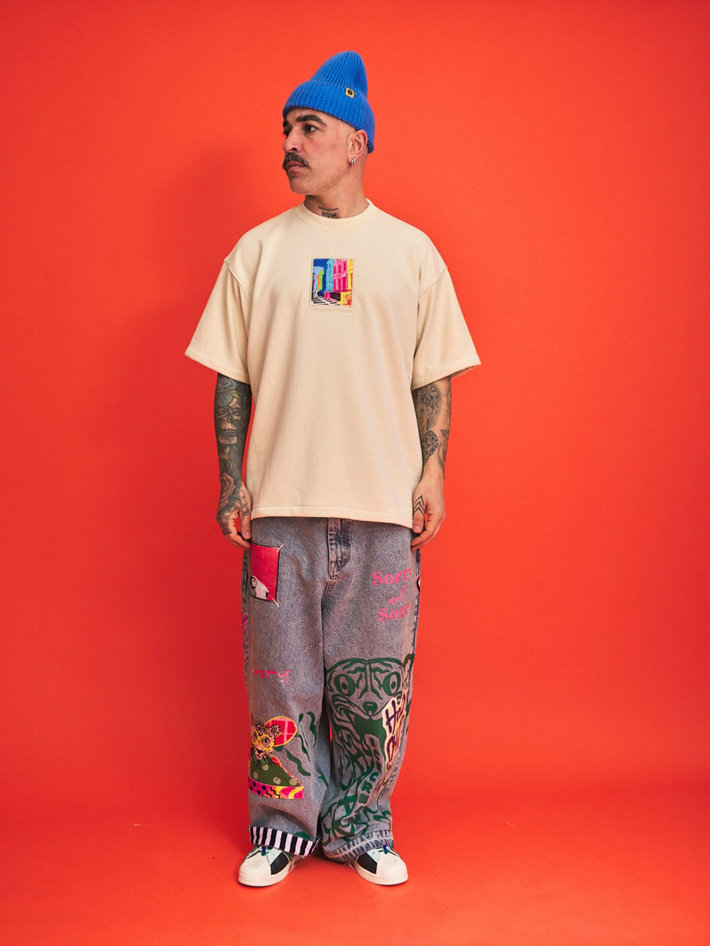 Man wearing a cream t-shirt, gray pants with designs, and a blue beanie on an orange background