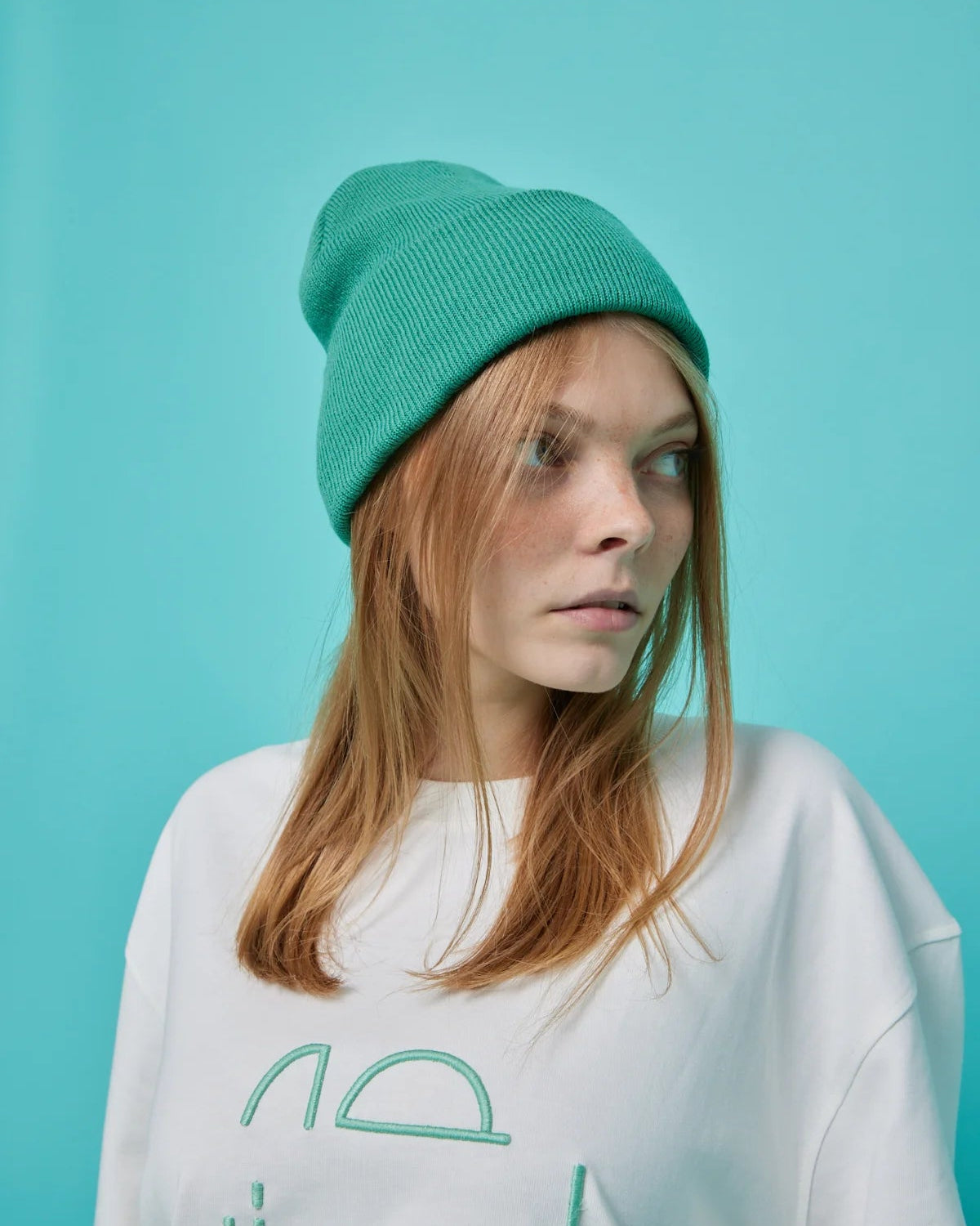 Person wearing a white sweatshirt with green text and a green beanie against a teal background