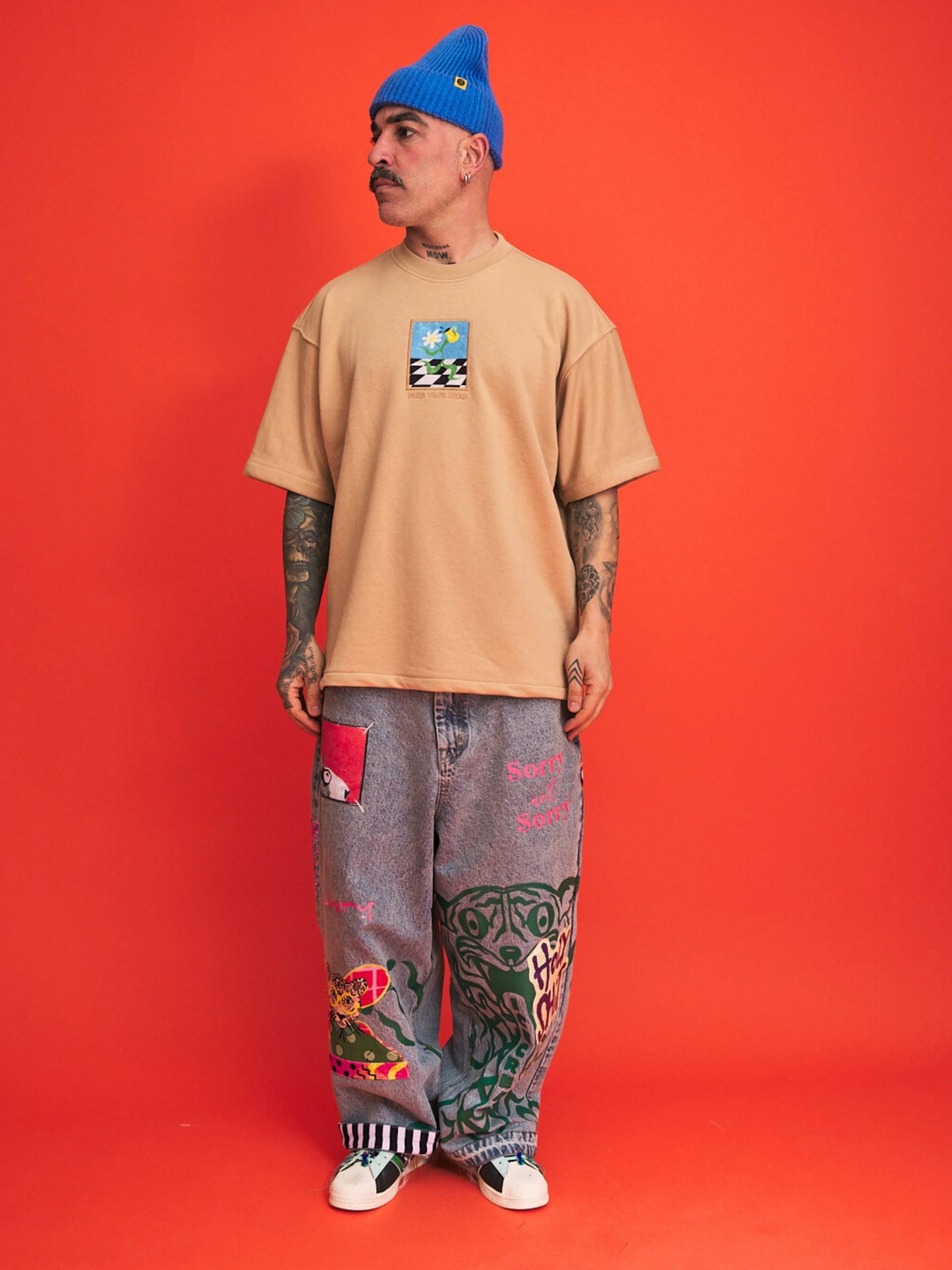 Person wearing a beige t-shirt with a graphic design and gray pants with colorful patches on an orange background