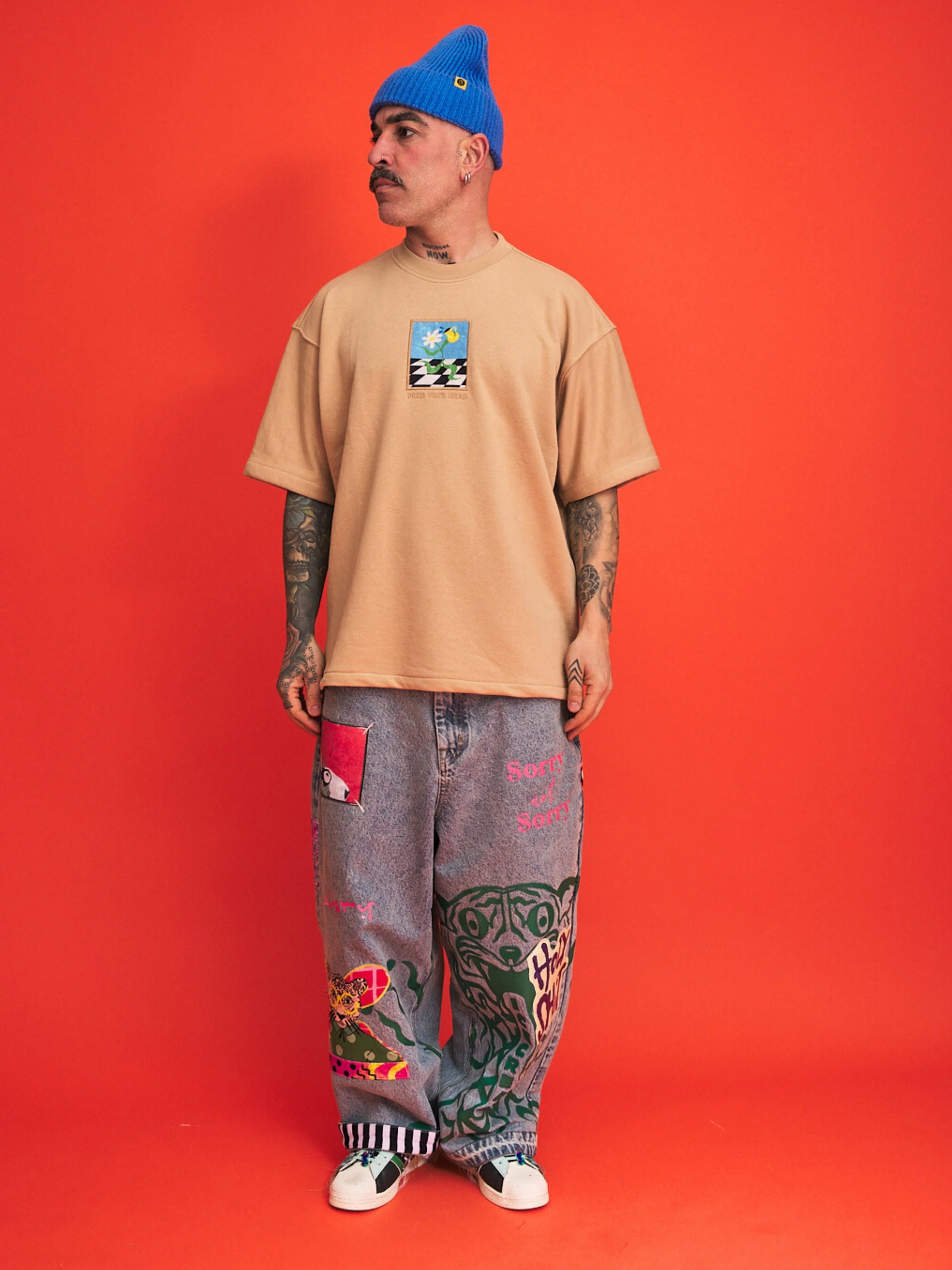 Person wearing a beige t-shirt with a graphic design and gray pants with colorful patches on an orange background