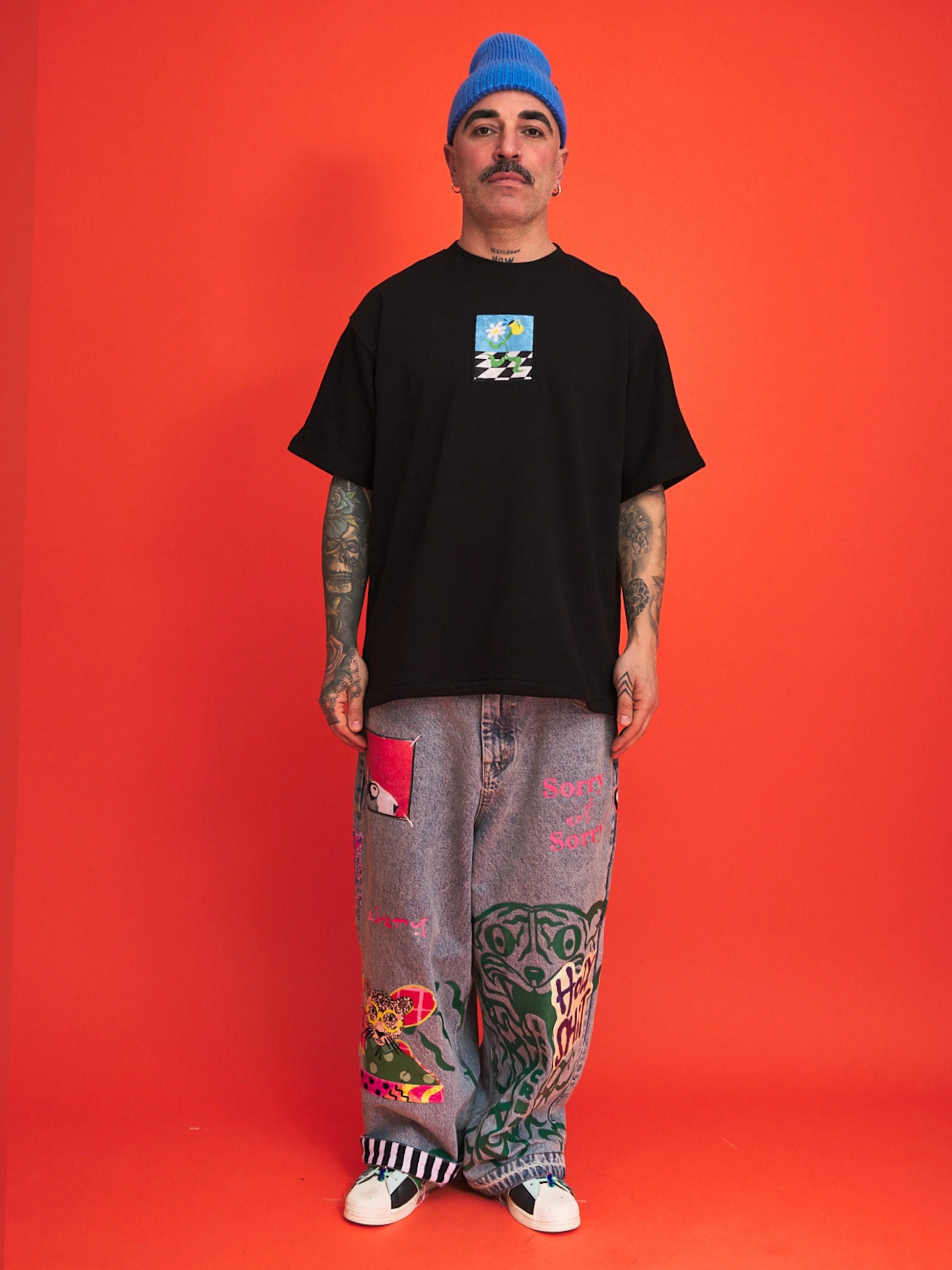 Person wearing a black t-shirt with a graphic design, gray pants with colorful patterns, and a blue beanie against an orange background.