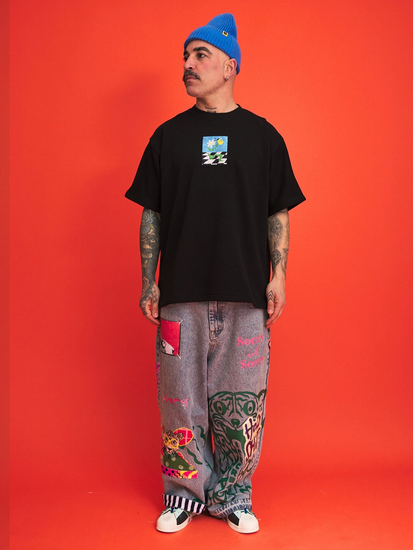 Person wearing a black t-shirt with a graphic design, colorful pants, and a blue beanie against an orange background