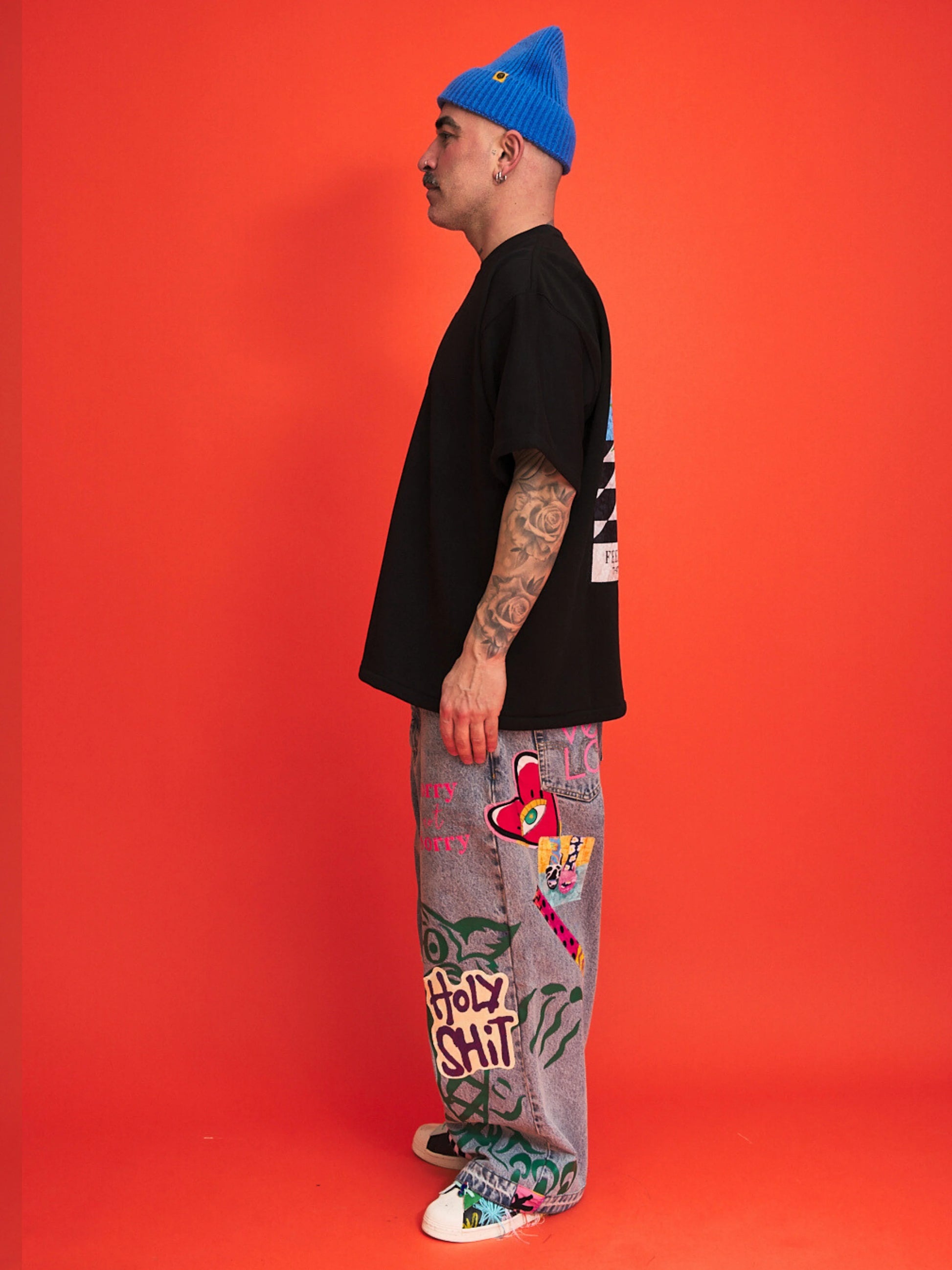 Person wearing a black t-shirt, colorful pants, and a blue beanie on an orange background