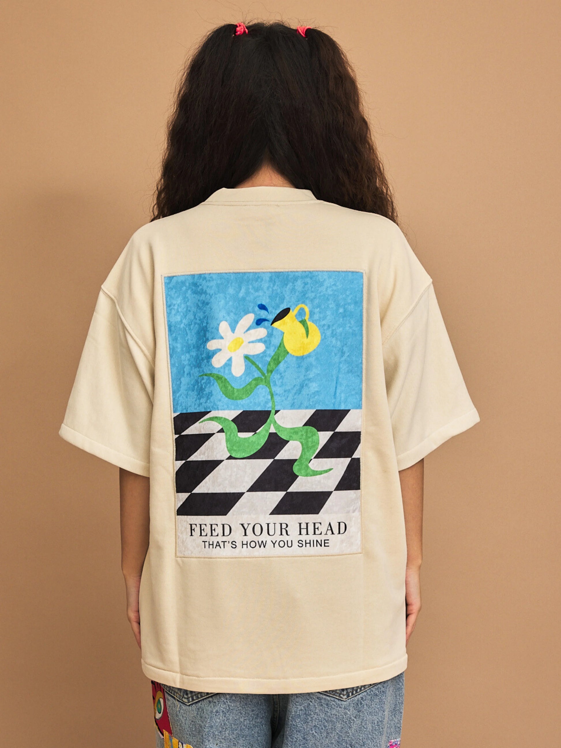 Person wearing a beige cream t-shirt with a graphic design on the back against a brown background