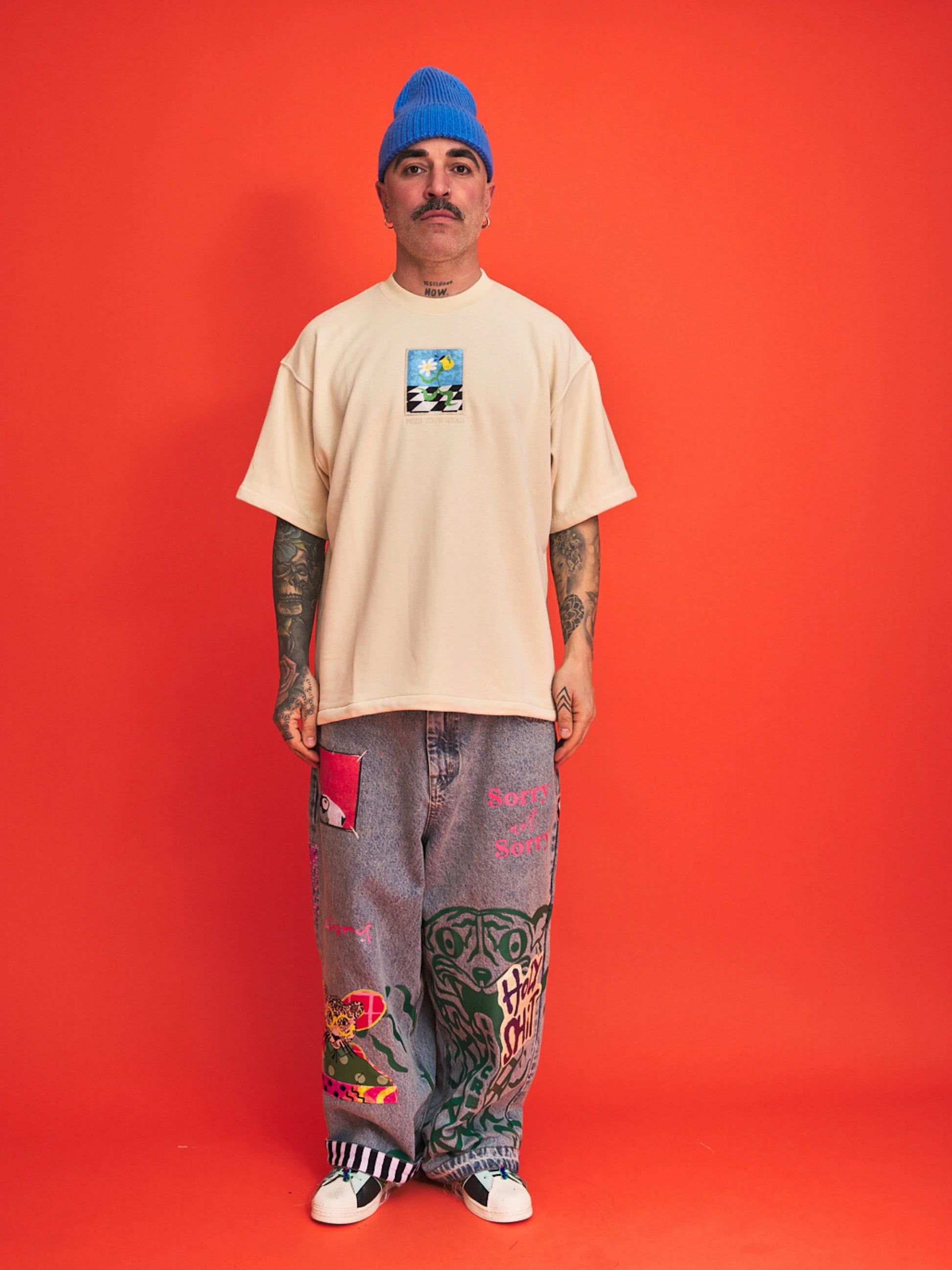 Person wearing a cream t-shirt, blue beanie, and patterned pants on an orange background