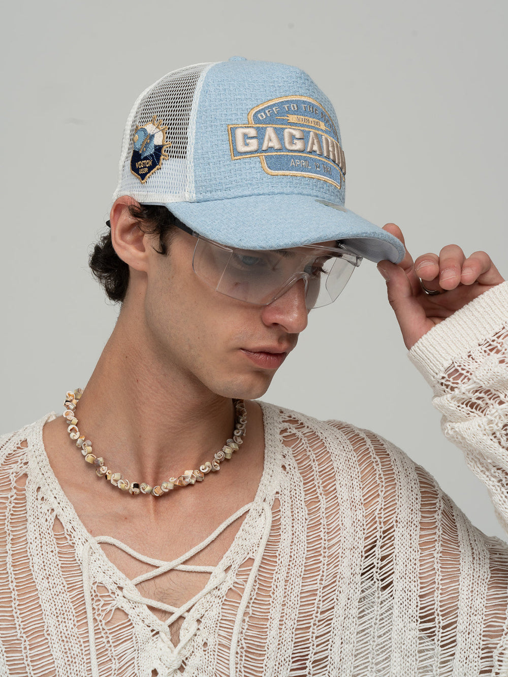 Person wearing Gagarin Blue Trucker Hat, white sweater, and necklace on a gray background