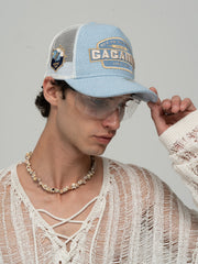 Person wearing Gagarin Blue Trucker Hat, white sweater, and necklace on a gray background