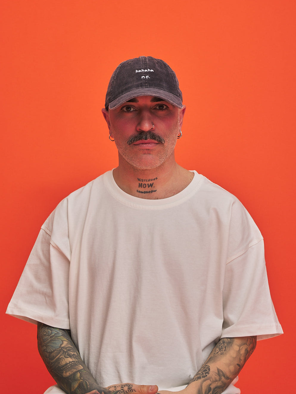 Man wearing a charcoal grey corduroy cap and white t-shirt against an orange background