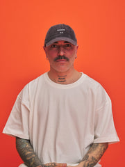 Man wearing a charcoal grey corduroy cap and white t-shirt against an orange background