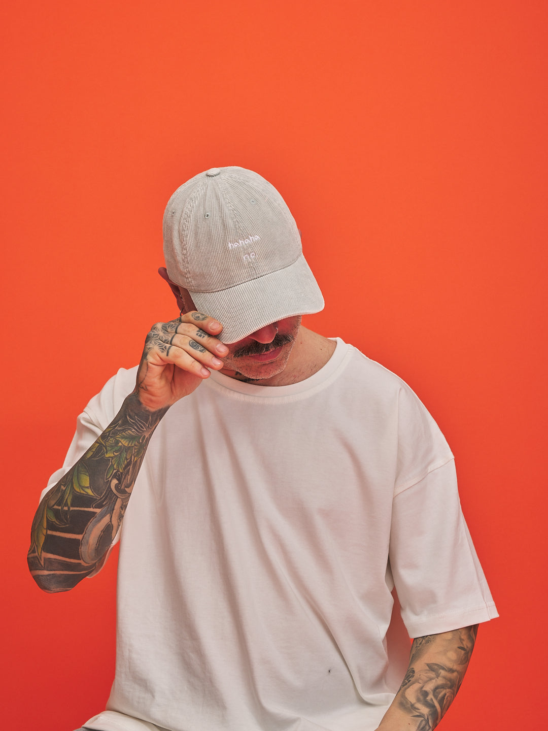 Person wearing a Hahaha No Light Grey Corduroy Cap and white t-shirt against an orange background