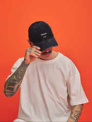 Person wearing a navy blue corduroy cap and white t-shirt against an orange background