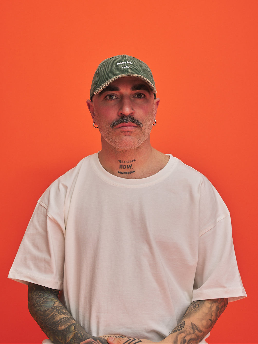 Man wearing a olive green corduroy cap and white t-shirt against an orange background