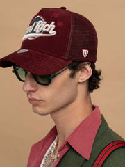Person wearing Hard Rich Burgundy Trucker Hat and sunglasses on a beige background