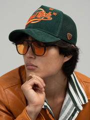 Person wearing Hard Rich Green Trucker Hat, sunglasses, and a brown leather jacket.
