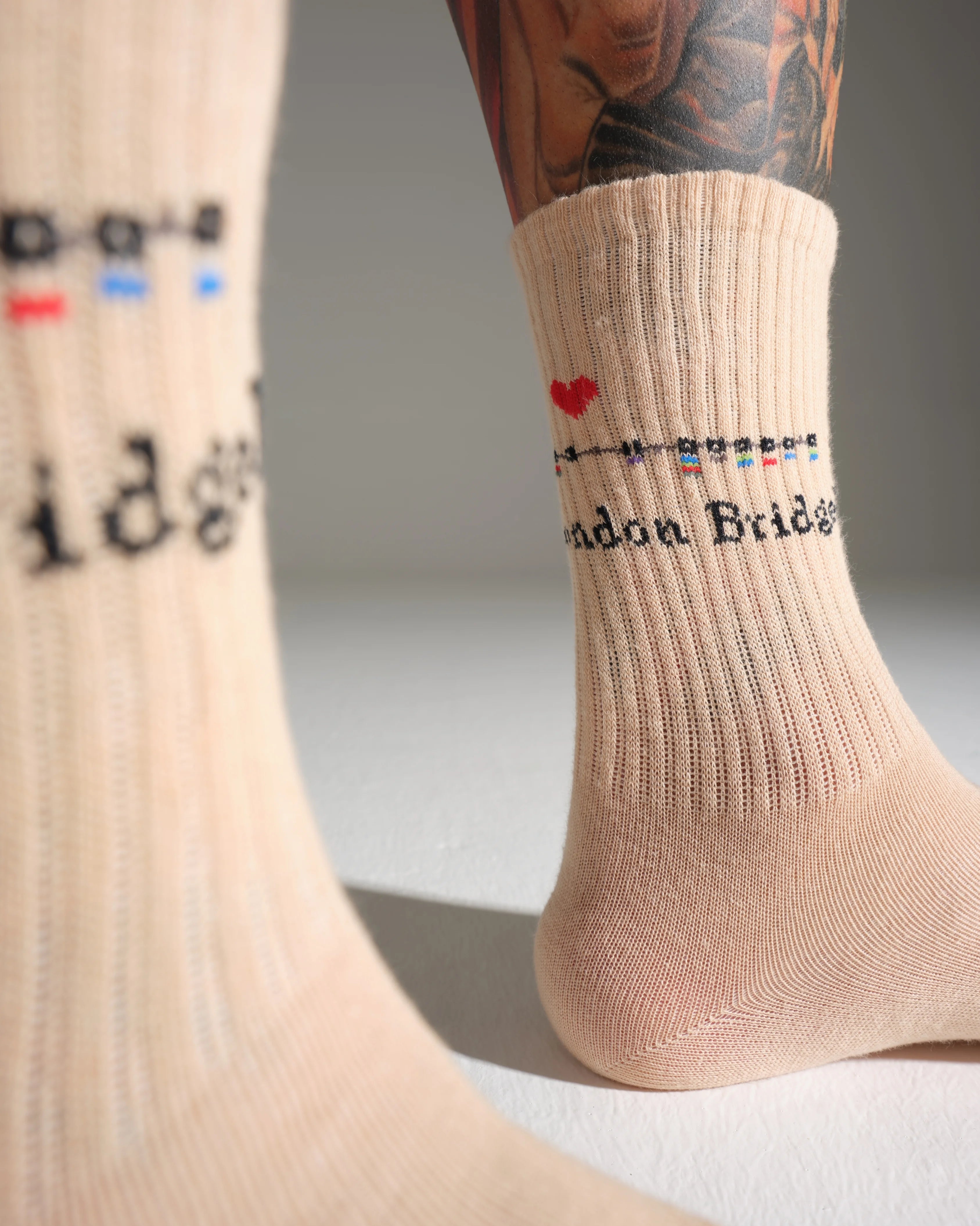 Beige London Bridge location and text socks with a heart design worn by a person with tattoos.
