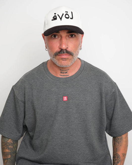 front look of man wearing a gray t-shirt with a small red logo and a white baseball cap with an black brim and Arabic 'love' text on a plain background.