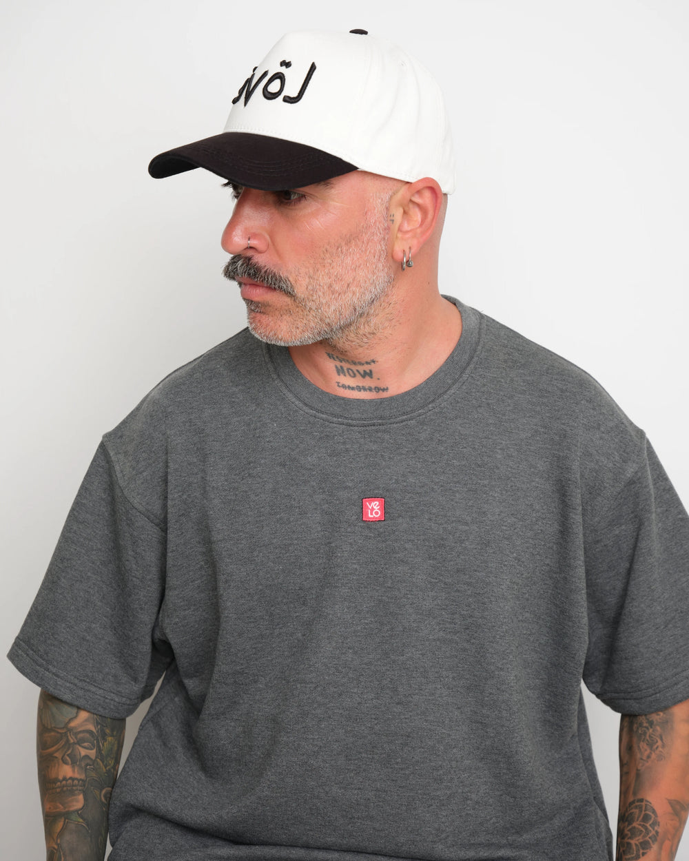 side look of man wearing a gray t-shirt with a small red logo and a white baseball cap with an black brim and Arabic 'love' text on a plain background.