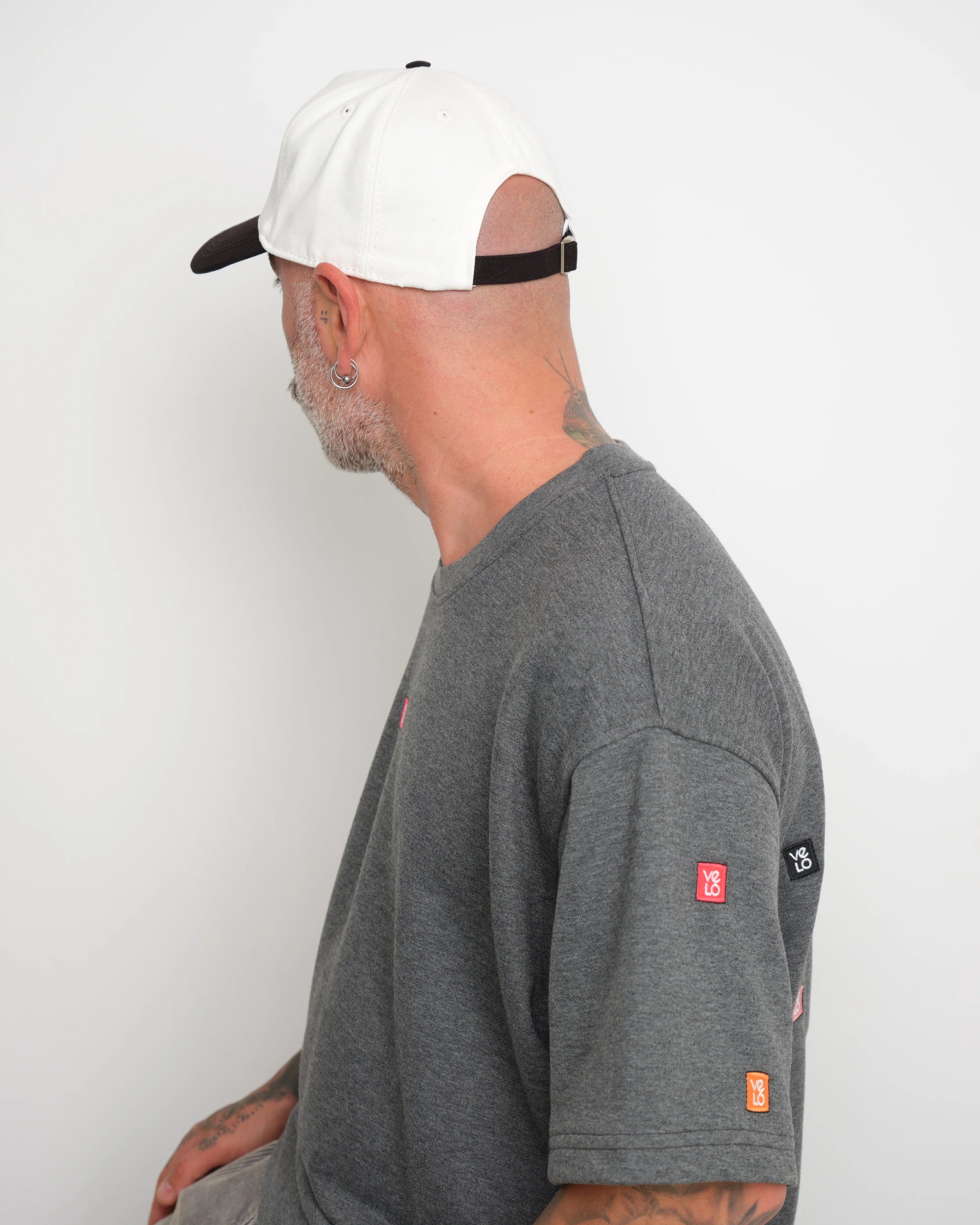 Man wearing a gray t-shirt with visible brand logos on a white background and showing brown flip tag of the cap 