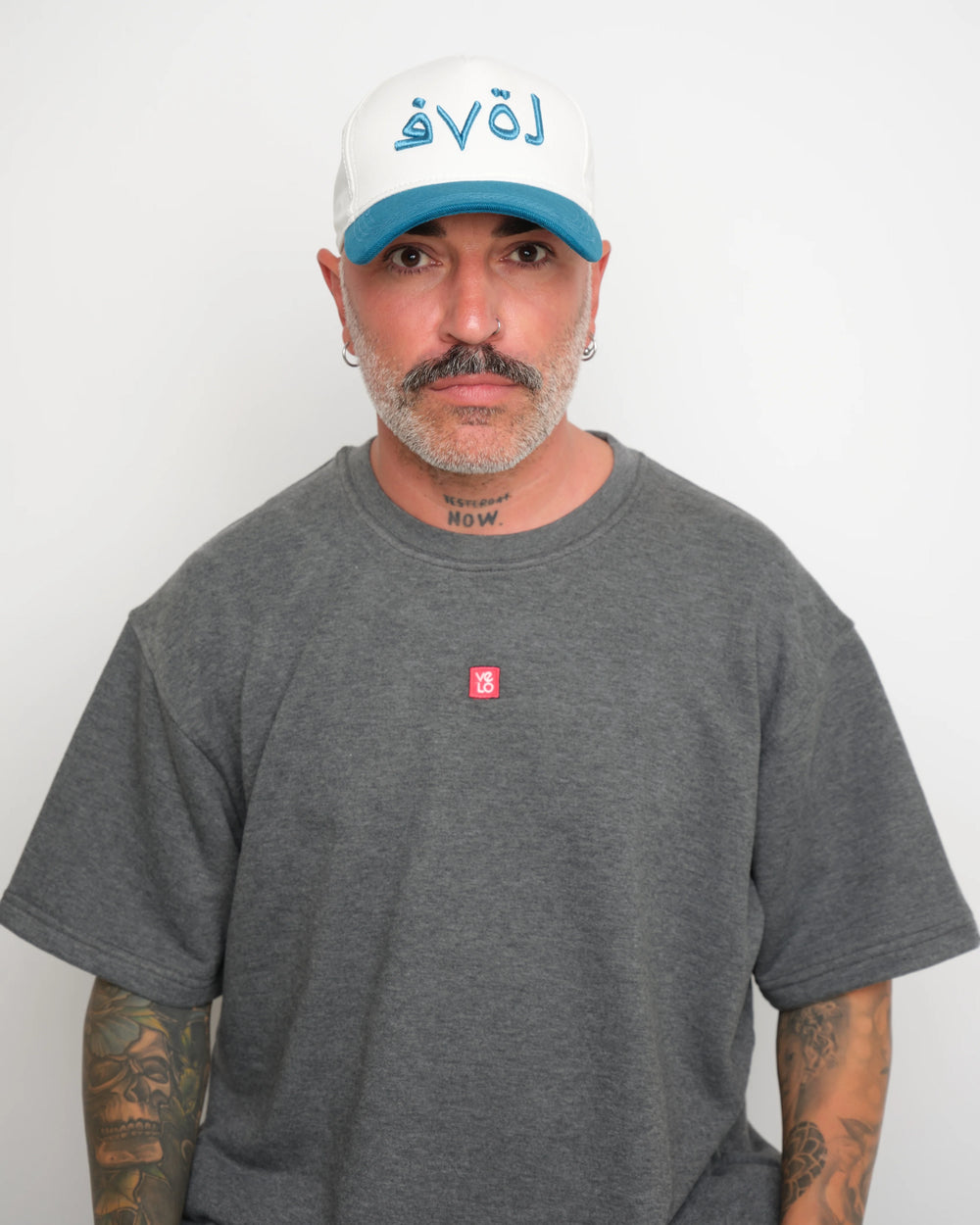 Man wearing a gray t-shirt with a small red VELO logo and a white cap with blue brim and blue Arabic 'love' text on a plain background