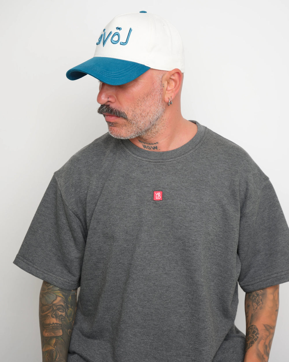 side look of man wearing a gray t-shirt with a small red VELO logo and a white cap with blue brim and blue Arabic 'love' text on a plain background
