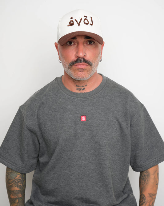 Man wearing a gray t-shirt with a small red logo and a white baseball cap with an brown brim and Arabic 'love' text on a plain background.