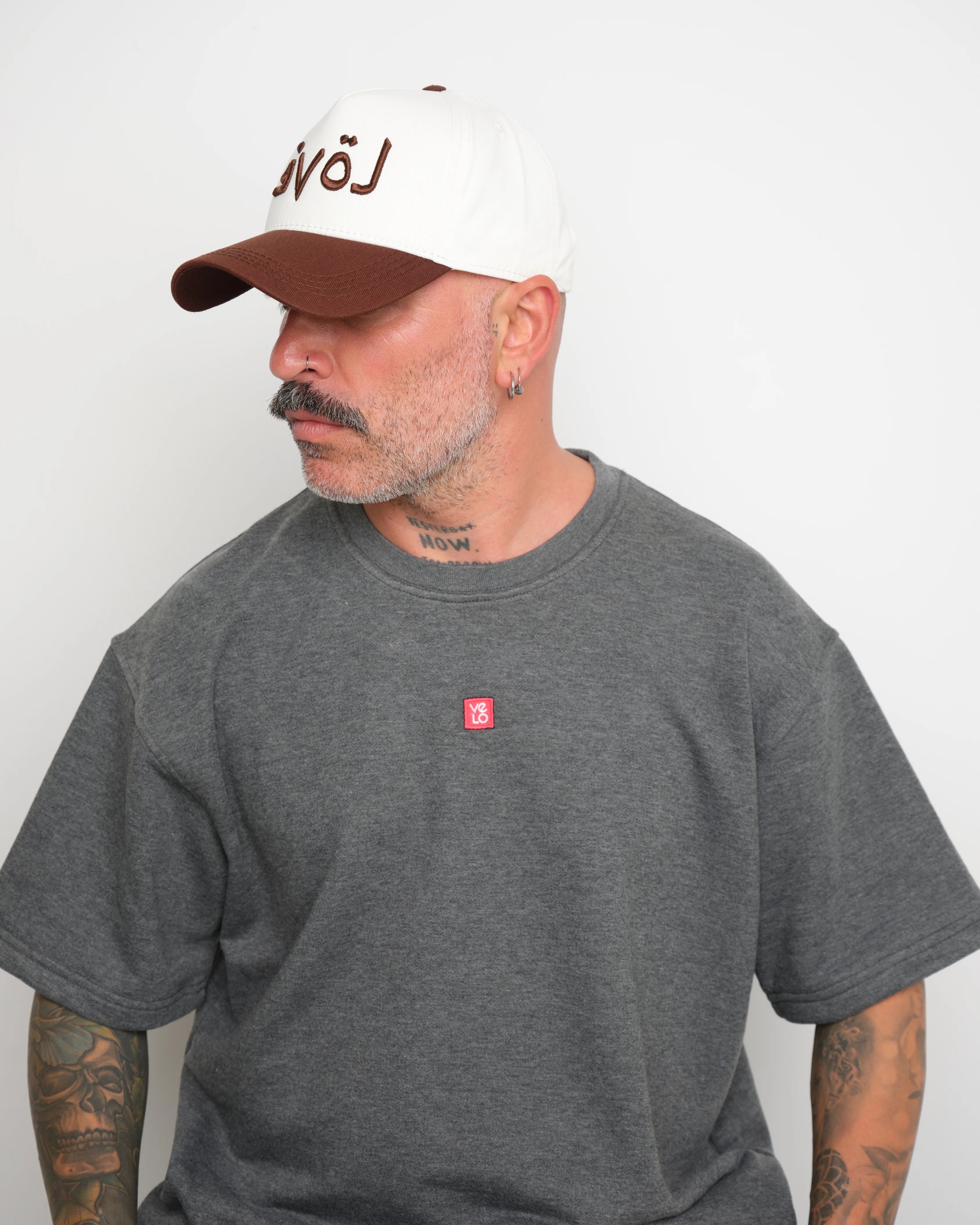 side look of man wearing a gray t-shirt with a small red logo and a white baseball cap with an brown brim and Arabic 'love' text on a plain background.