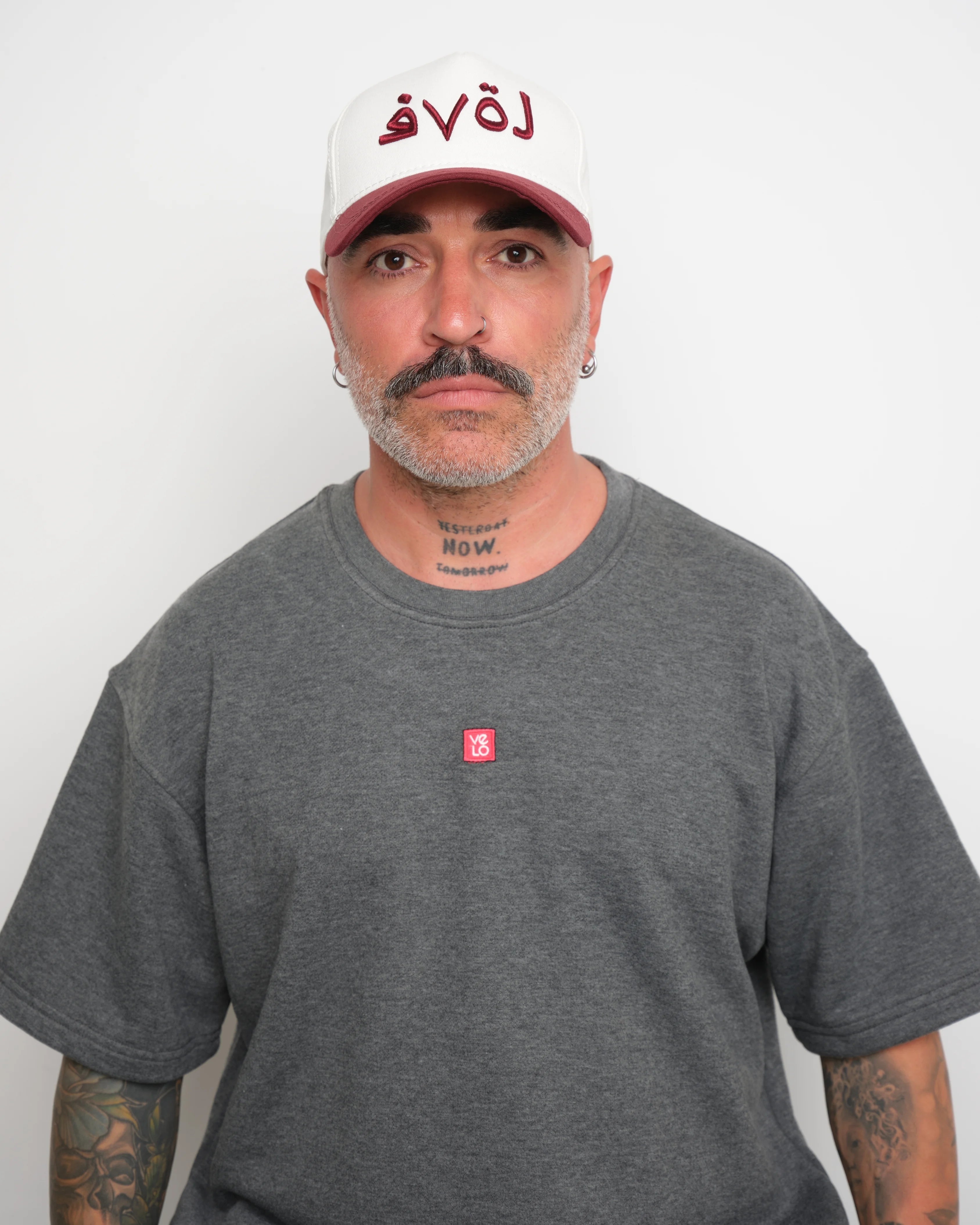 Man wearing a gray t-shirt with a small red logo and a white baseball cap with an burgundy brim and Arabic 'love' text on a plain background.