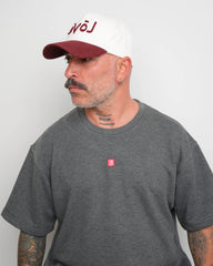 side look of man wearing a gray t-shirt with a small red logo and a white baseball cap with an burgundy brim and Arabic 'love' text on a plain background.