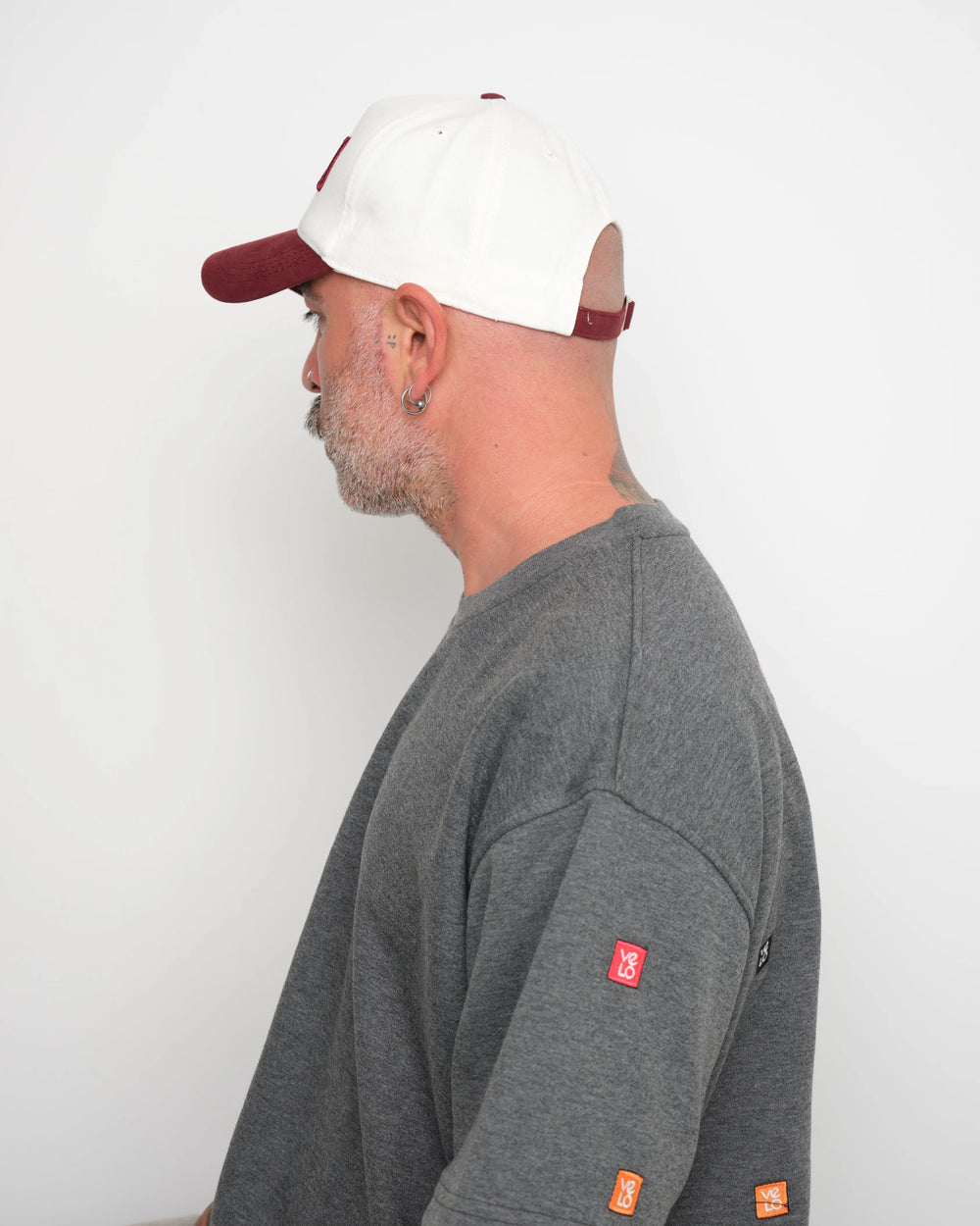 Man wearing a gray t-shirt with 'velo' geometric stitched and showing flip tag of baseball cap on a white background