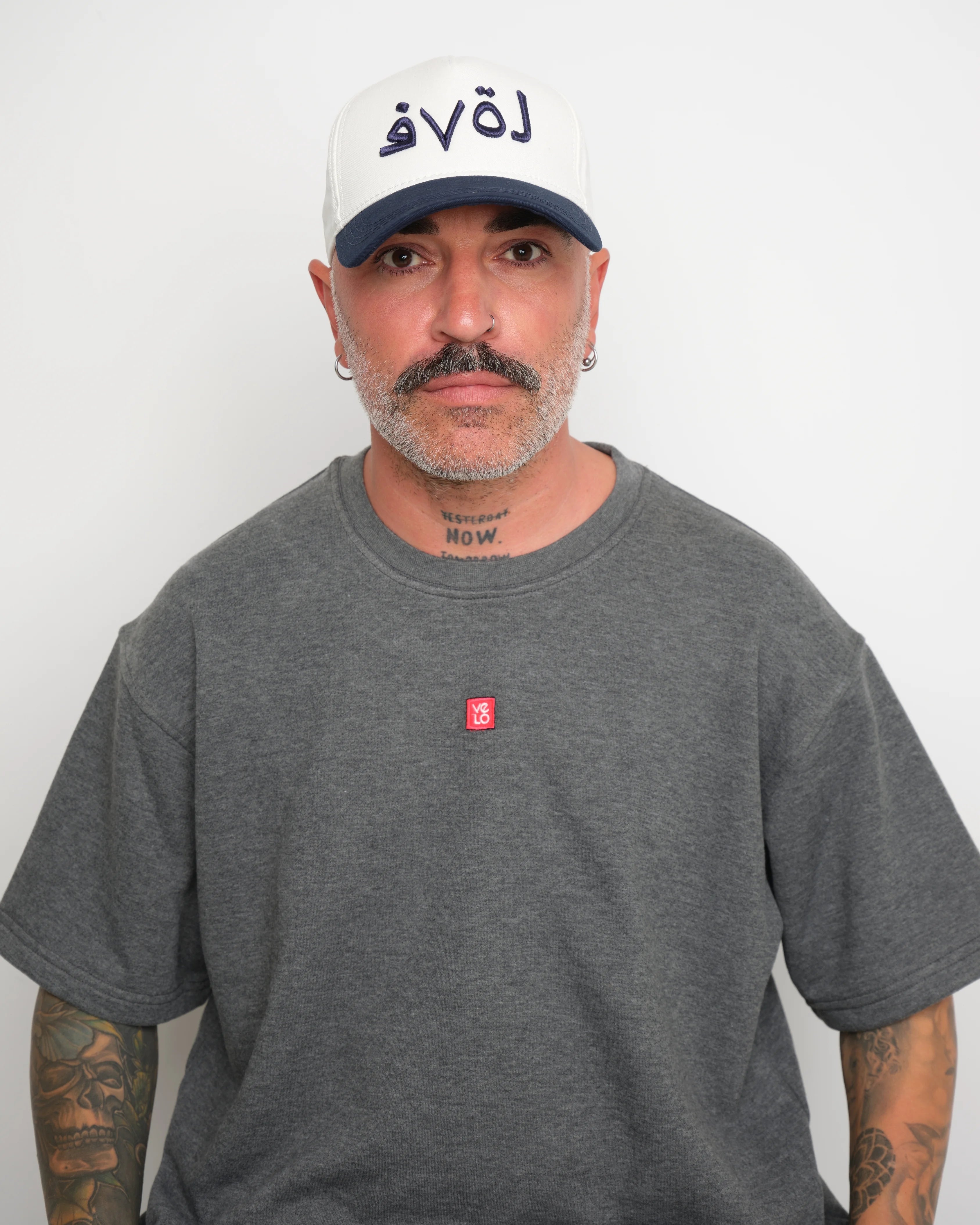 Man wearing a gray t-shirt with a small red logo and a white baseball cap with an dark navy brim and Arabic 'love' text on a plain background.