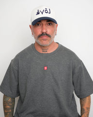 Man wearing a gray t-shirt with a small red logo and a white baseball cap with an dark navy brim and Arabic 'love' text on a plain background.