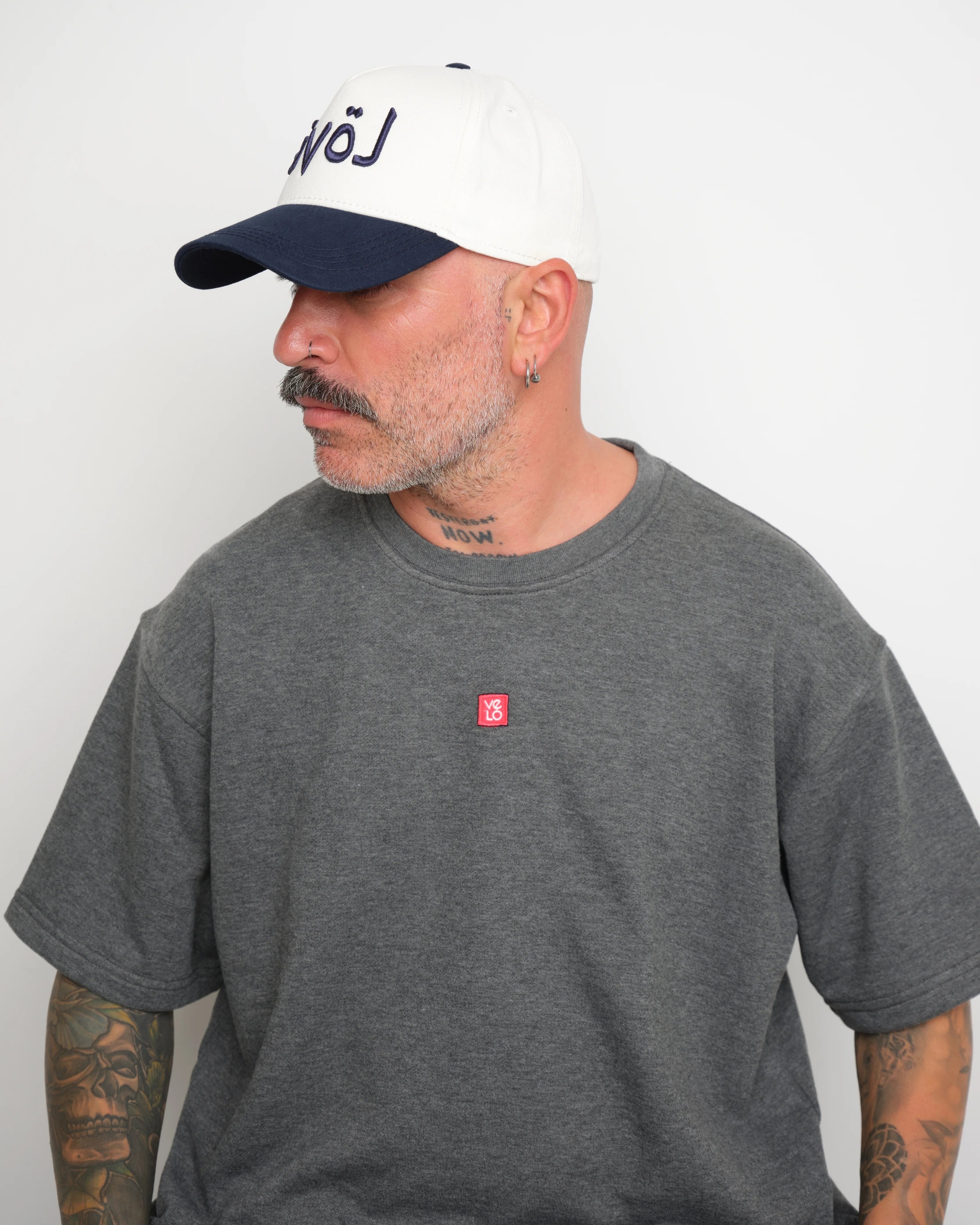 Side look of man wearing a gray t-shirt with a small red logo and a white baseball cap with an dark navy brim and Arabic 'love' text on a plain background.