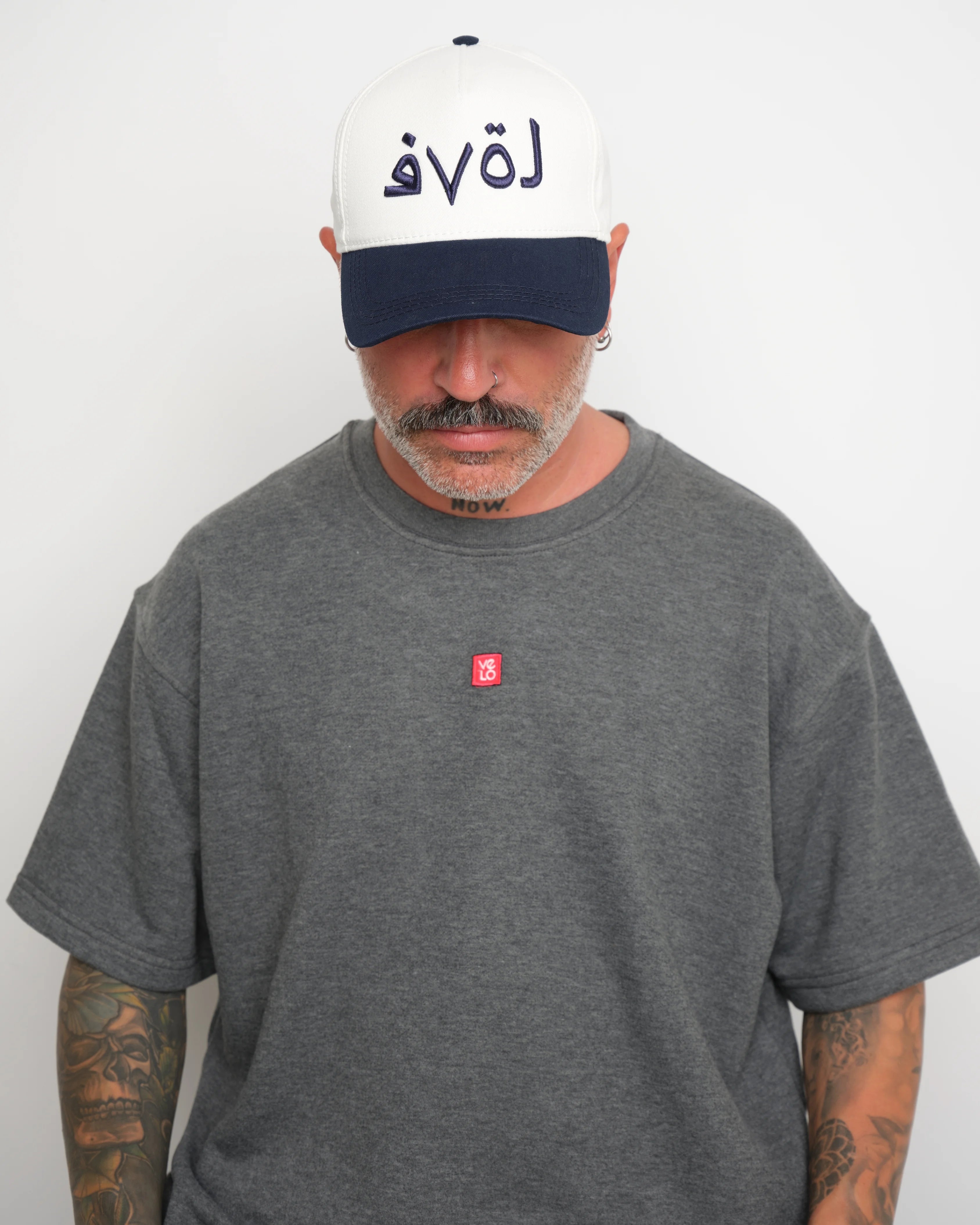 front look of man wearing a gray t-shirt with a small red logo and a white baseball cap with an dark navy brim and Arabic 'love' text on a plain background.