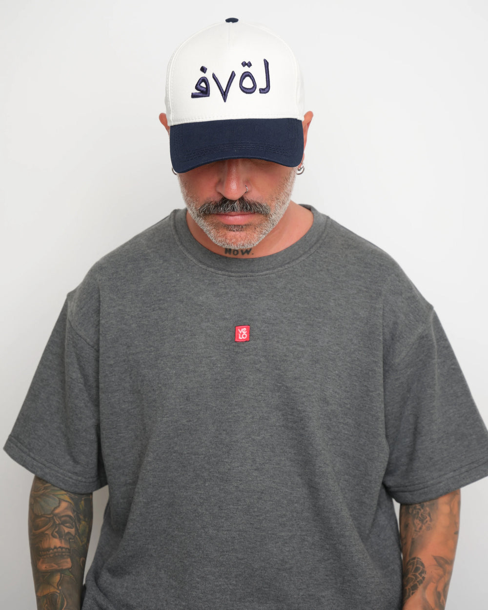 front look of man wearing a gray t-shirt with a small red logo and a white baseball cap with an dark navy brim and Arabic 'love' text on a plain background.