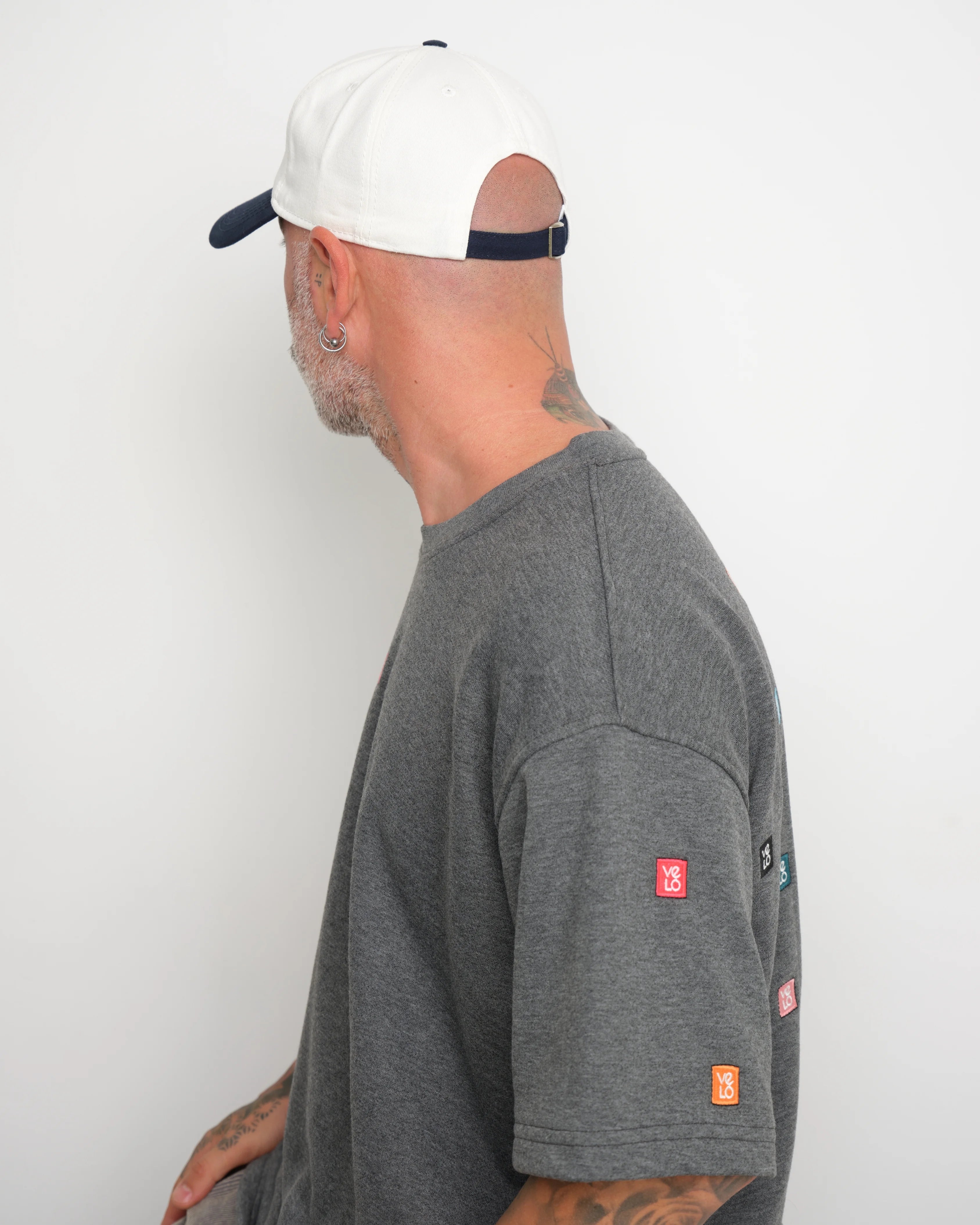 Man wearing a gray t-shirt with 'velo' geometric stitched and showing flip tag of baseball cap on a white background