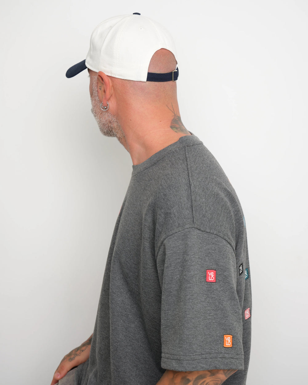 Man wearing a gray t-shirt with 'velo' geometric stitched and showing flip tag of baseball cap on a white background