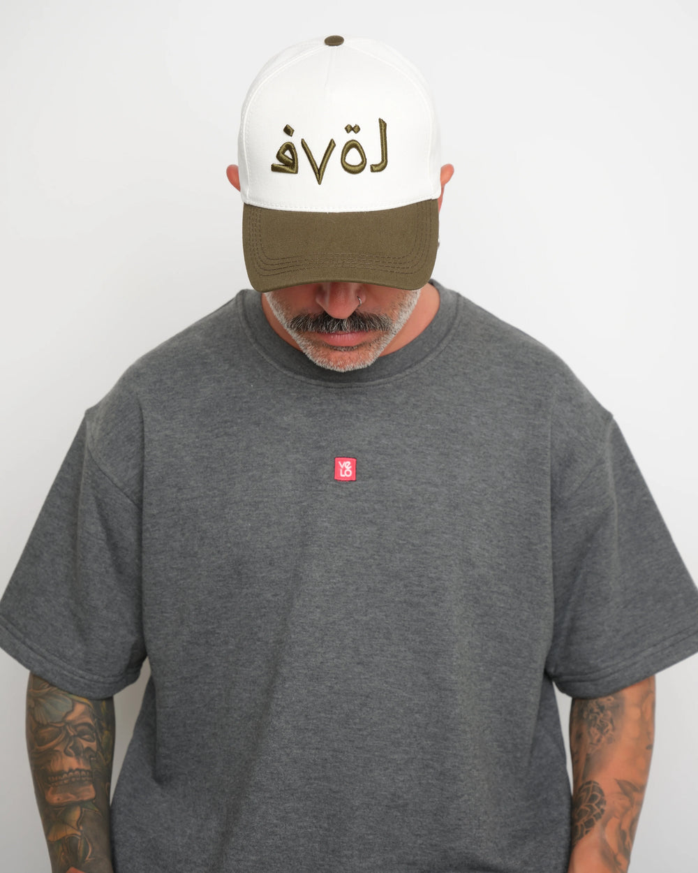 Man wearing a gray t-shirt with a red logo and a white cap with khaki brim and khaki Arabic 'love'  text on a plain background