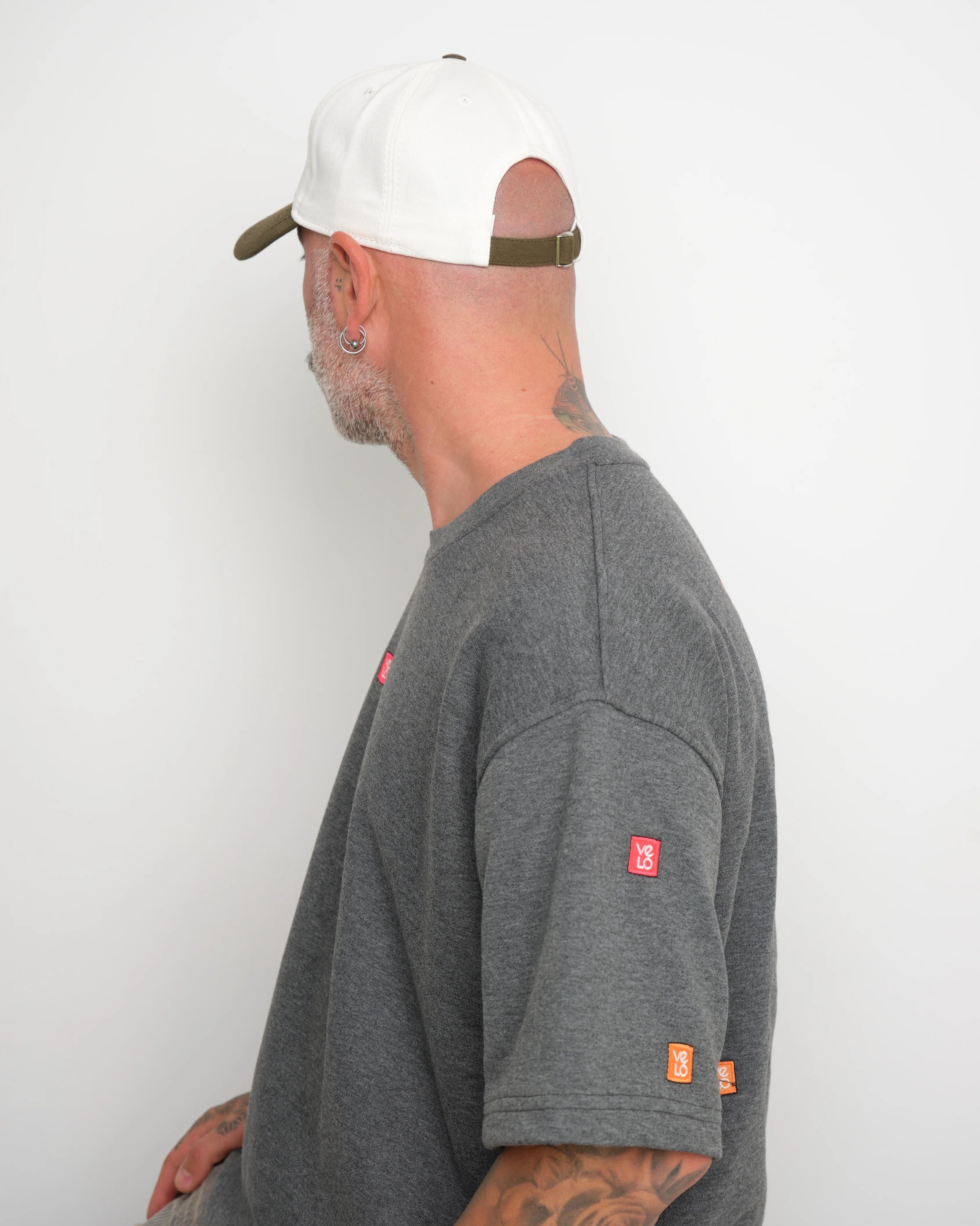 Man wearing a gray t-shirt with small logos on a white background and shows adjustable flip tag of the cap