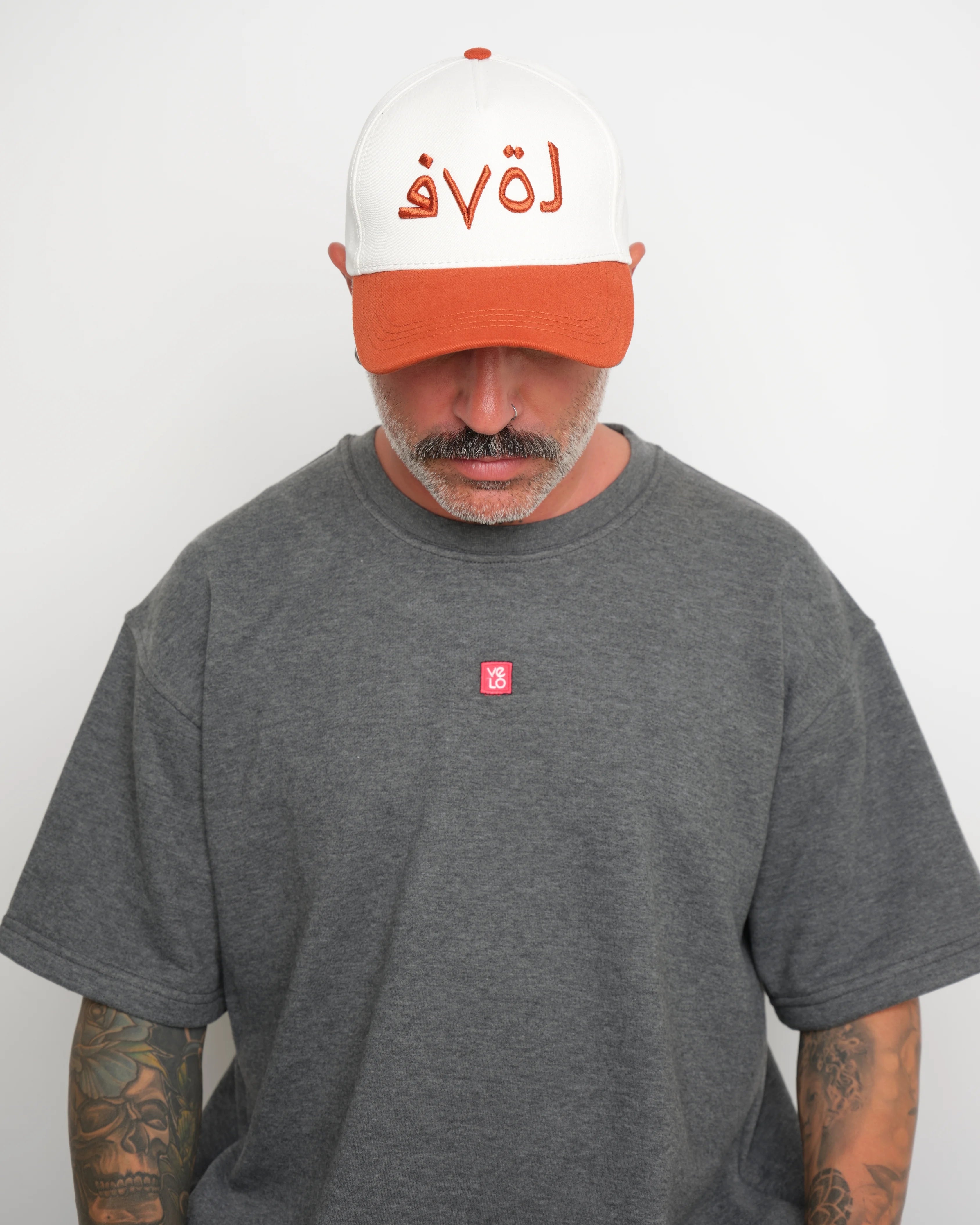Man wearing a gray t-shirt with a small red logo and a white baseball cap with an orange brim and orange Arabic text on a plain background.